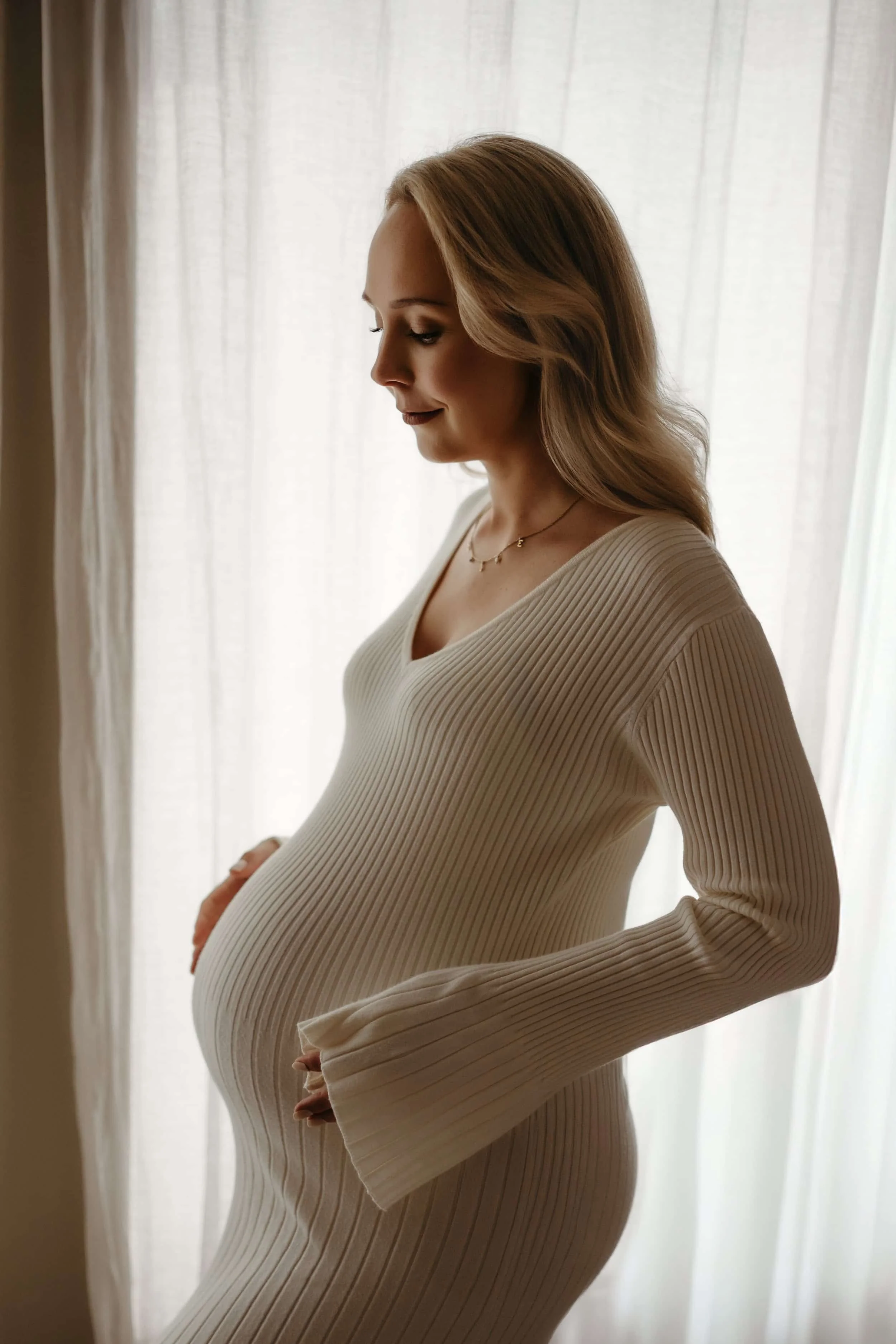 Profil av gravid kvinne med langt, lyst hår, iført en kremfarget, ribbet kjole, står foran et lyst gardin.
