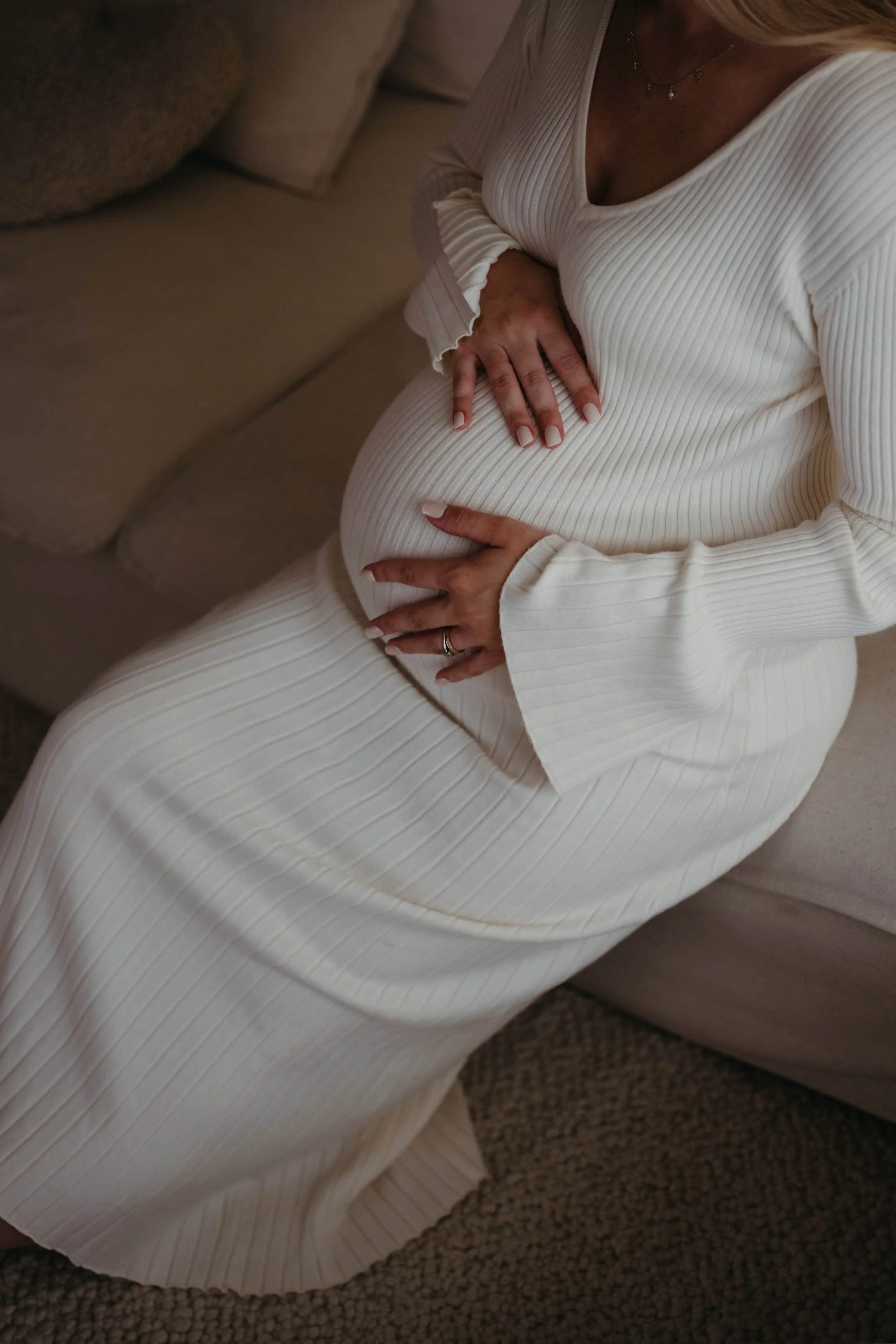 Nærbilde av en gravid kvinne som klemmer magen sin, iført en hvit ribbet kjole, sittende på et teppe omgitt av puter.