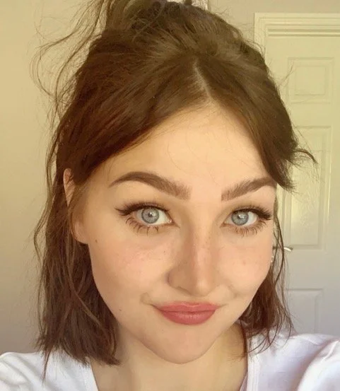 Close-up of a young woman with shoulder-length reddish-brown hair, blue eyes, and light makeup, standing indoors in front of a beige wall and a white door.