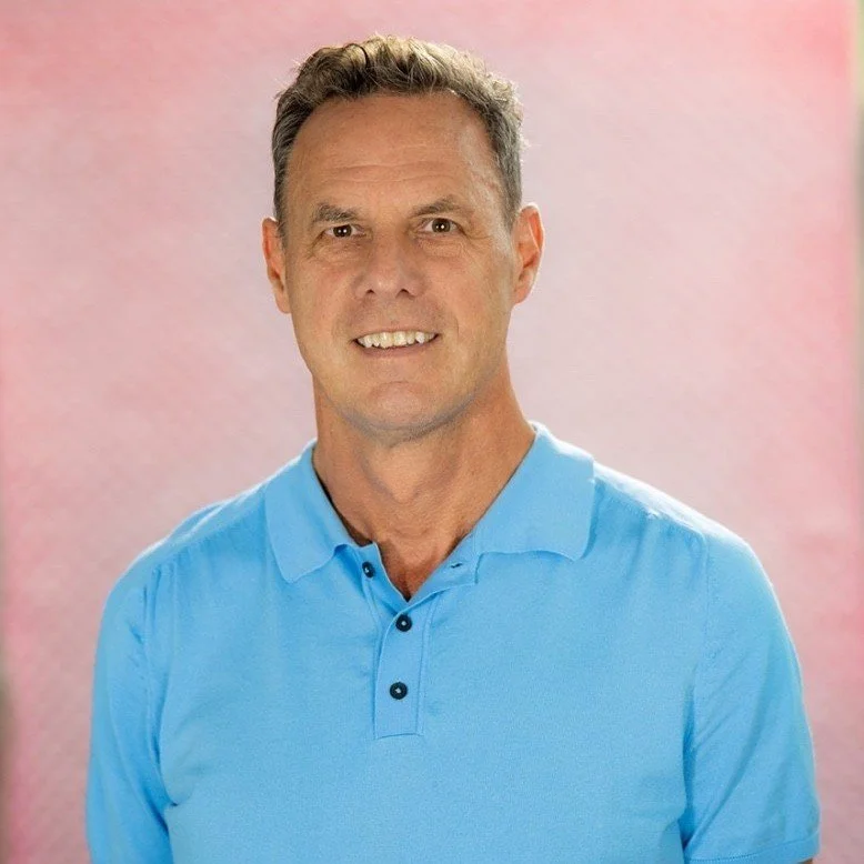Portrait of a middle-aged man with short gray hair, wearing a light blue polo shirt, smiling, against a pink textured background.