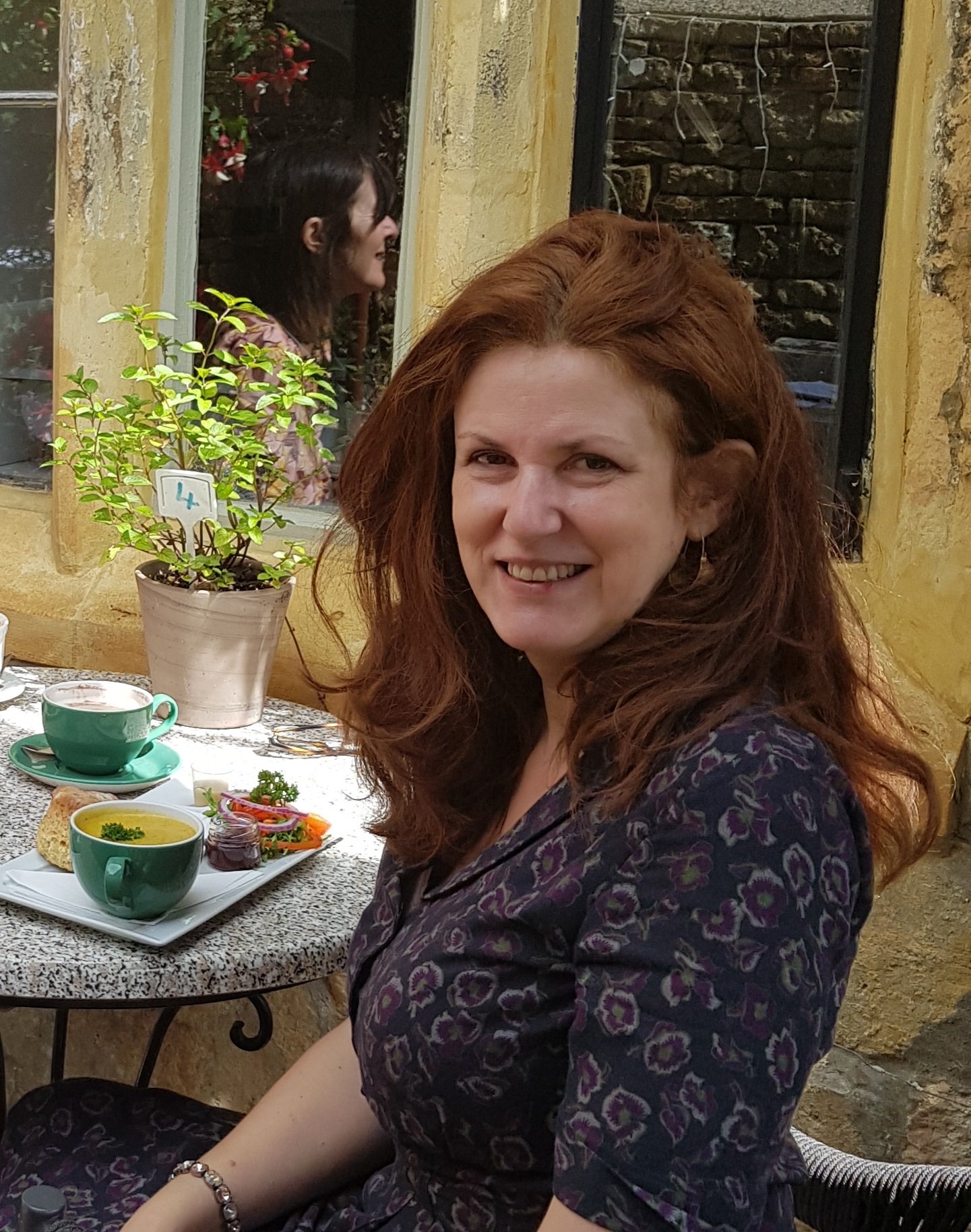 A woman with long red hair smiling at an outdoor cafe table, with soup, salad, and a cup of tea, near a yellow brick wall and window with a reflection of another woman.