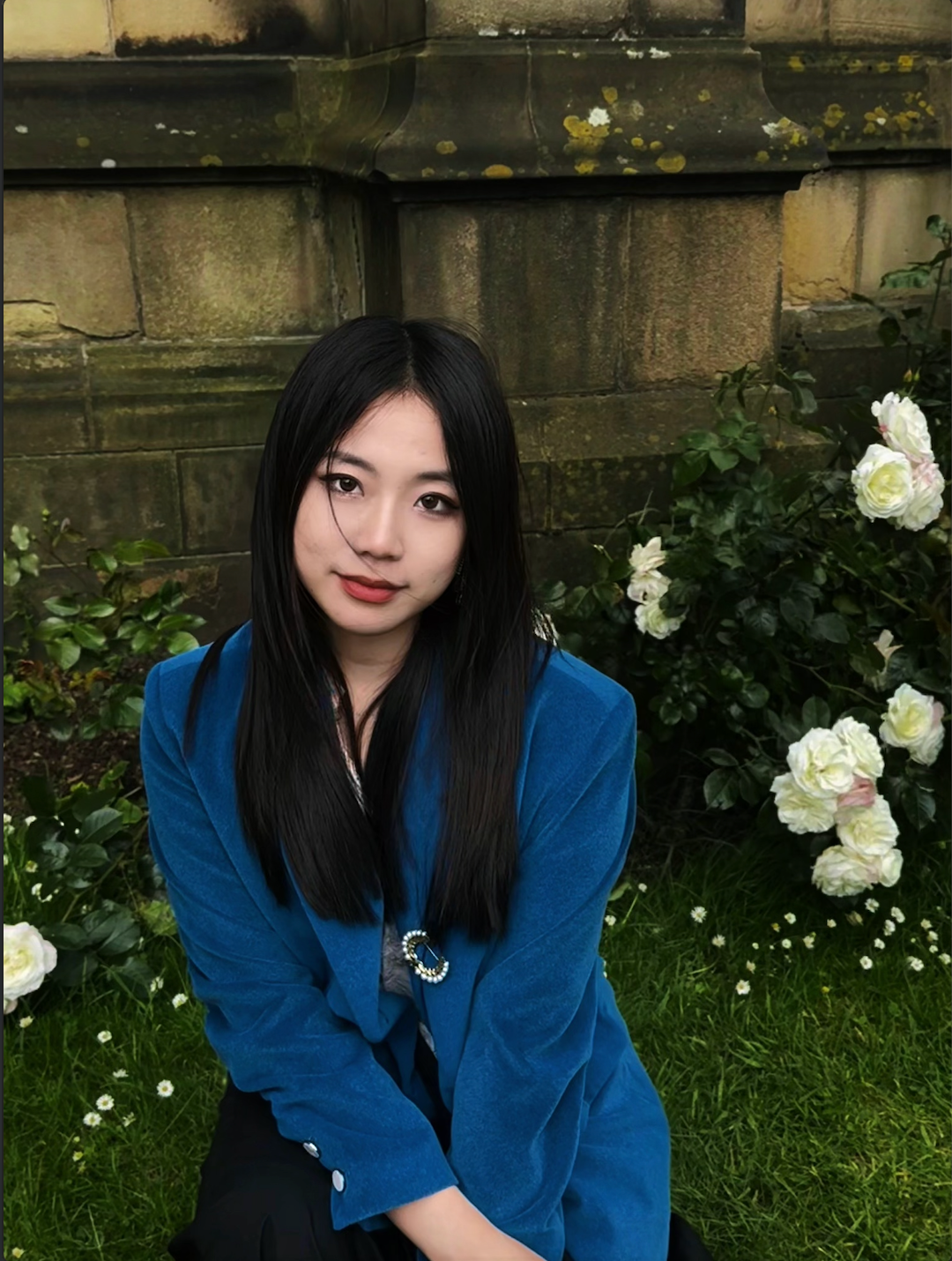 A young woman with straight black hair and light skin sits outdoors on grass, surrounded by white roses and green foliage, with an old stone building in the background.
