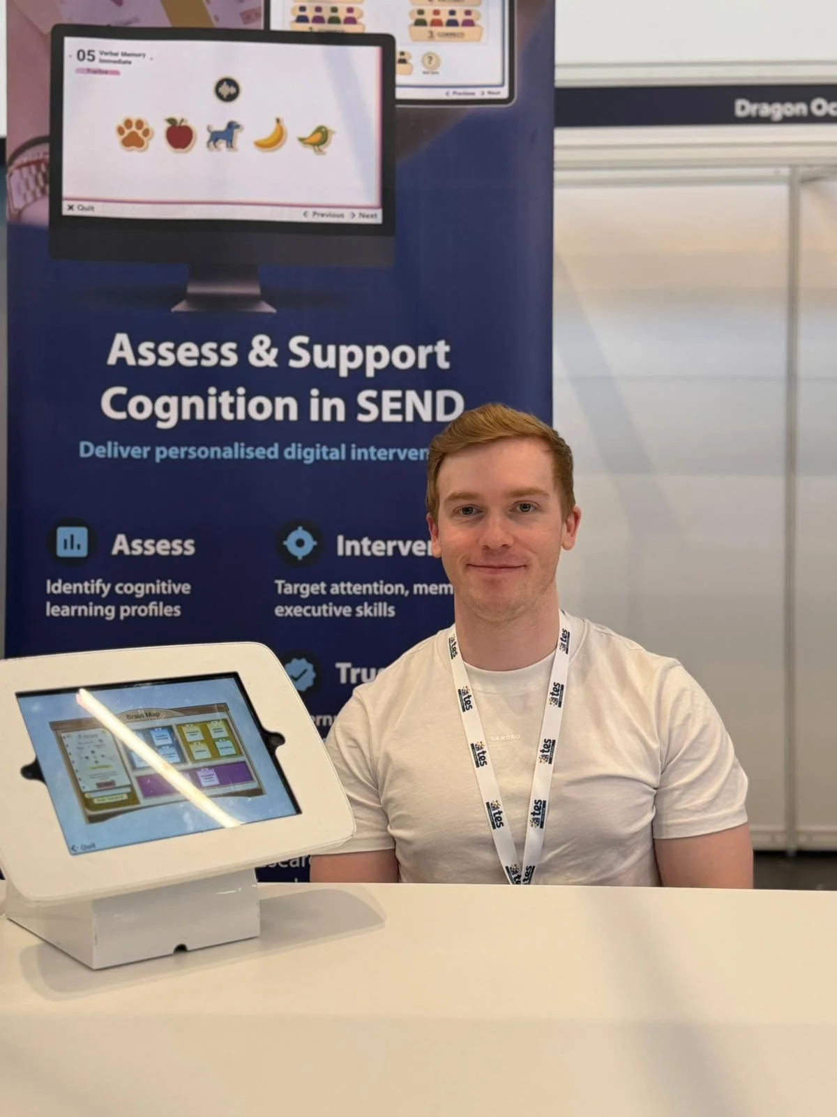 A young man with light skin, blonde hair, and a slight smile, sitting indoors near a window with a blurred cityscape in the background.