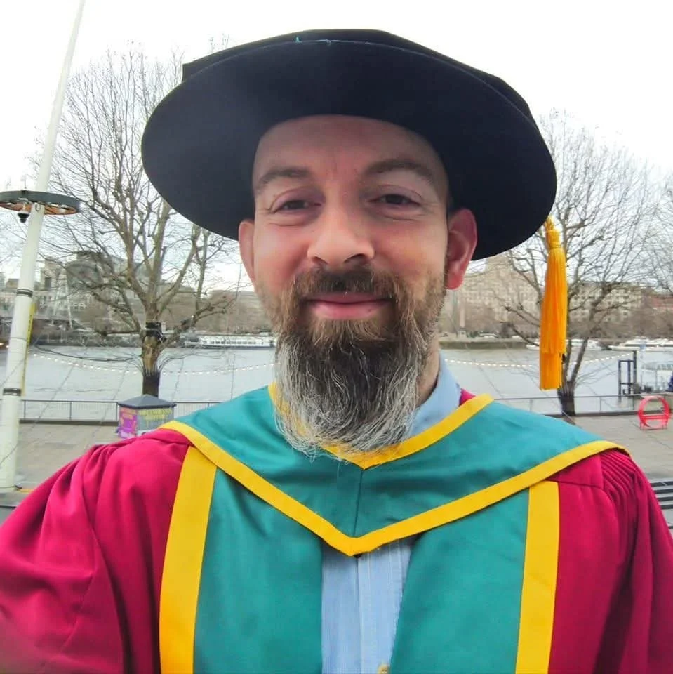 A man in graduation robes and cap smiling outdoors with trees and a body of water in the background.