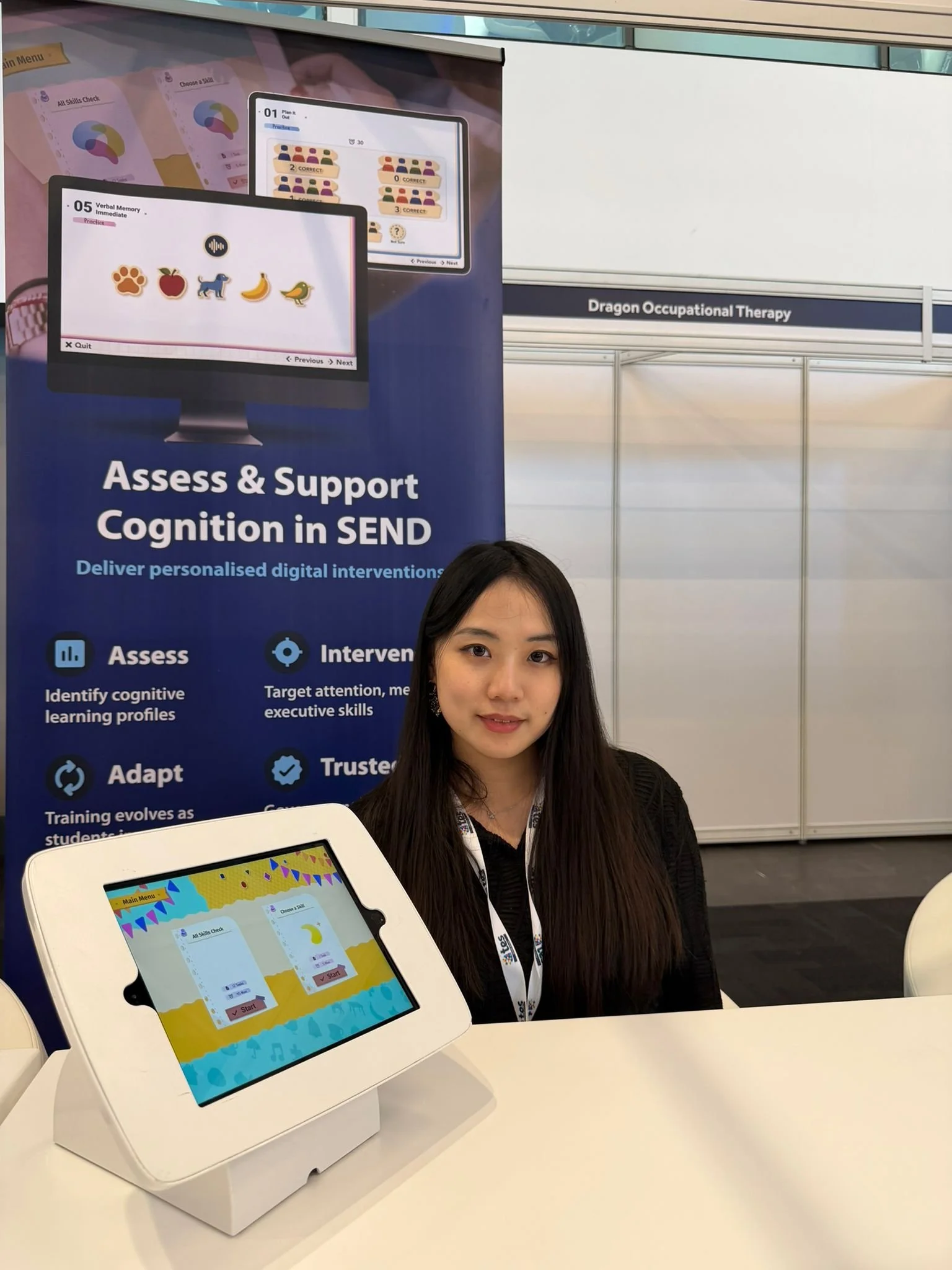 A young woman with long black hair and light skin, wearing a blue blazer with a decorative clasp, sitting outdoors on grass next to white flowers and green foliage, with an aged stone building in the background.