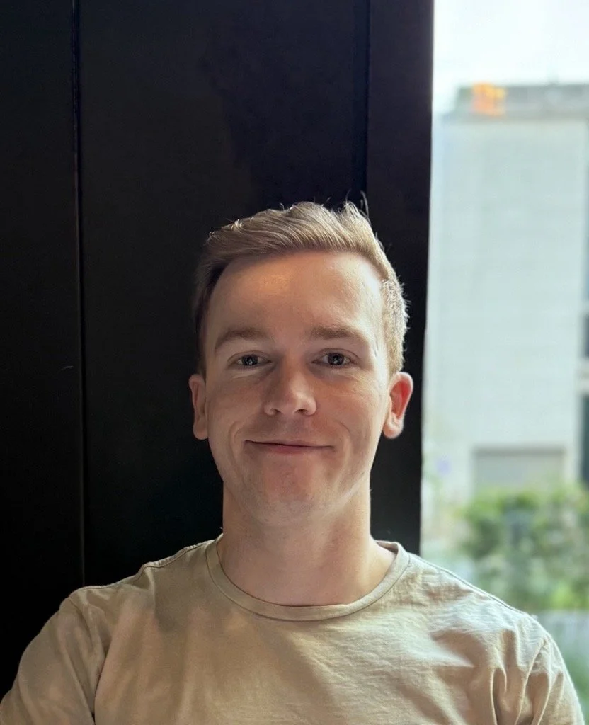 A young man with light skin, blonde hair, and a slight smile, sitting indoors near a window with a blurred cityscape in the background.