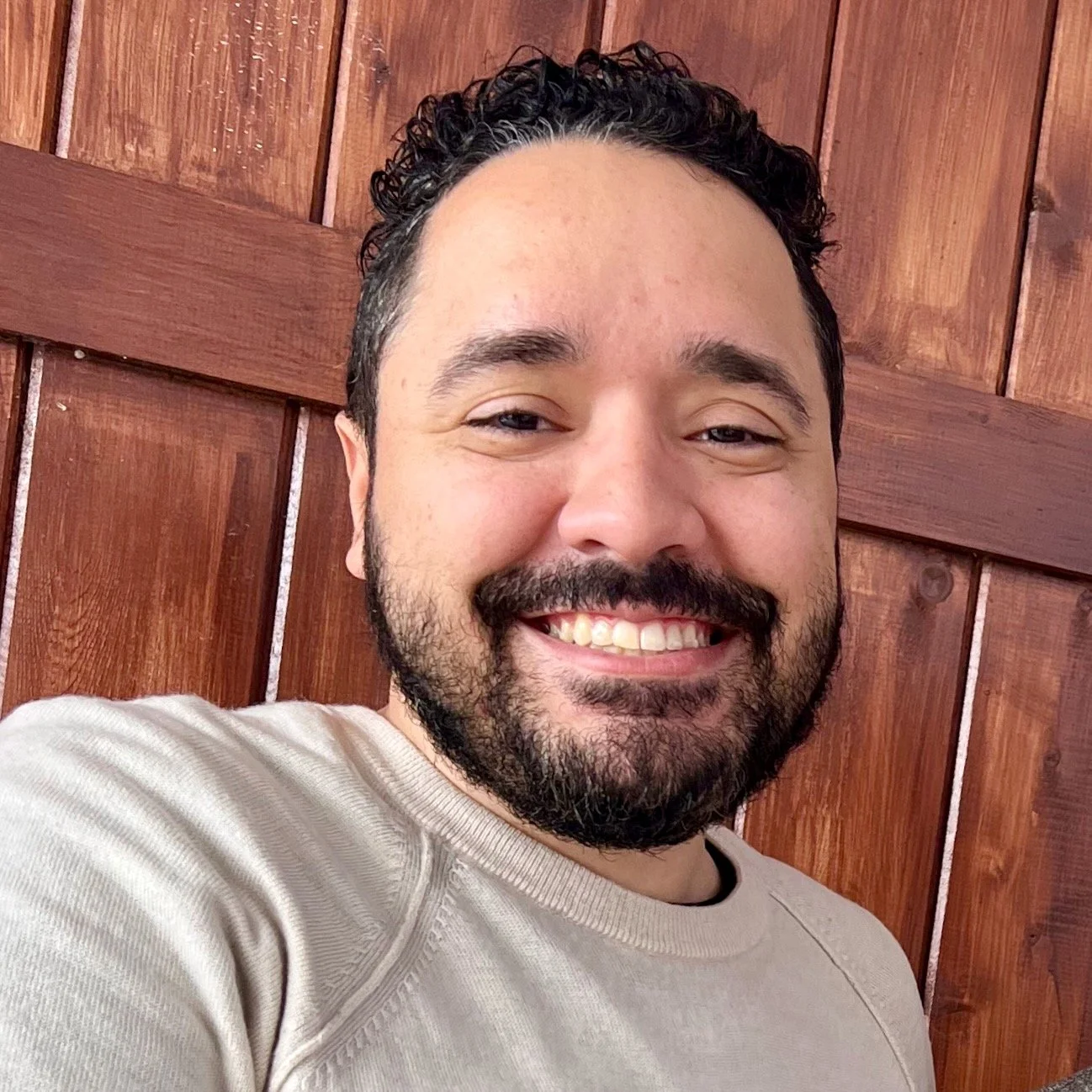 A smiling man with dark curly hair and a beard, wearing a beige shirt, standing in front of a wooden wall.