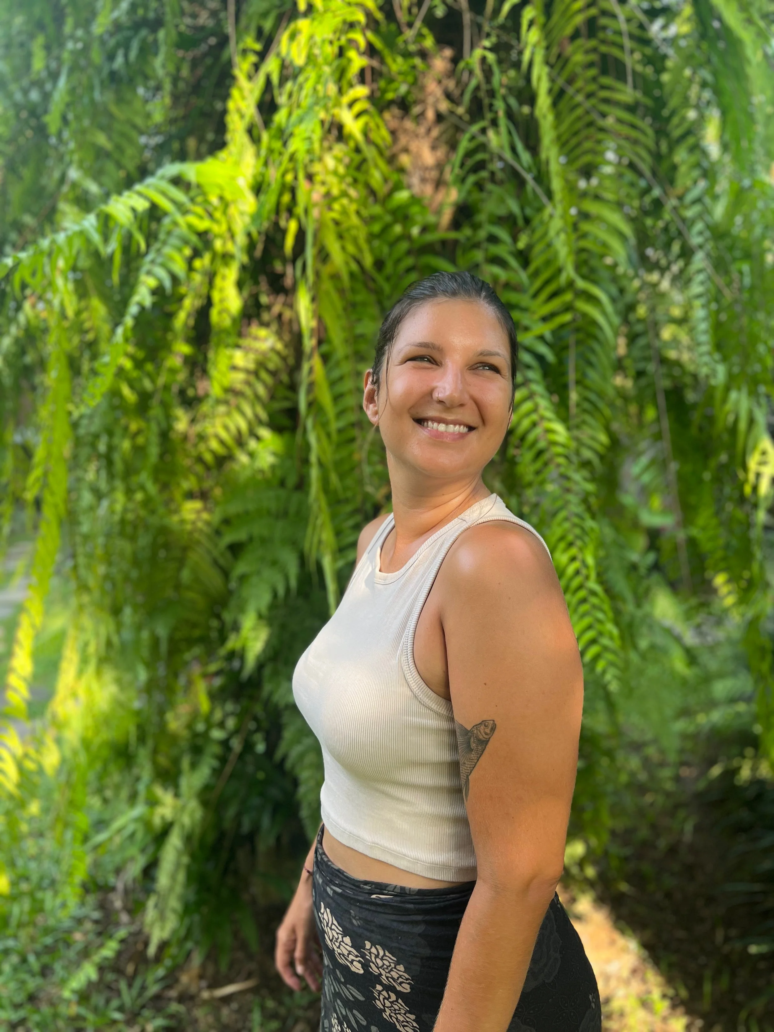 Eine lachende Frau in einem weißen Top und schwarzen Musterhose im grünen Dschungel.