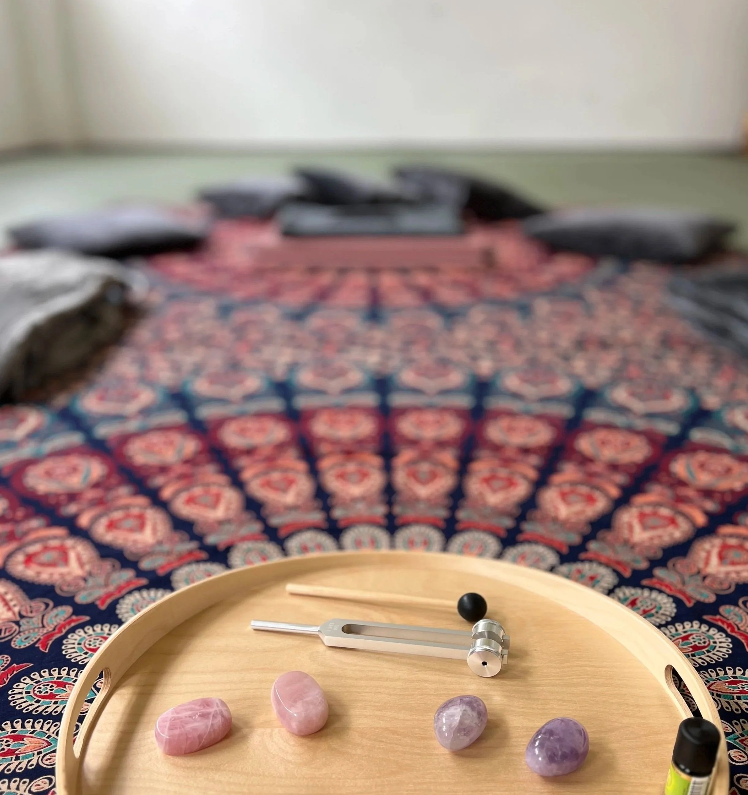 Rundes Holztablett mit Heilsteinen, einer Stimmgabel, ätherischen Ölen, auf einem bunten Mandala-Tuch, einer Yoga-Matte mit mehreren Kissen.
