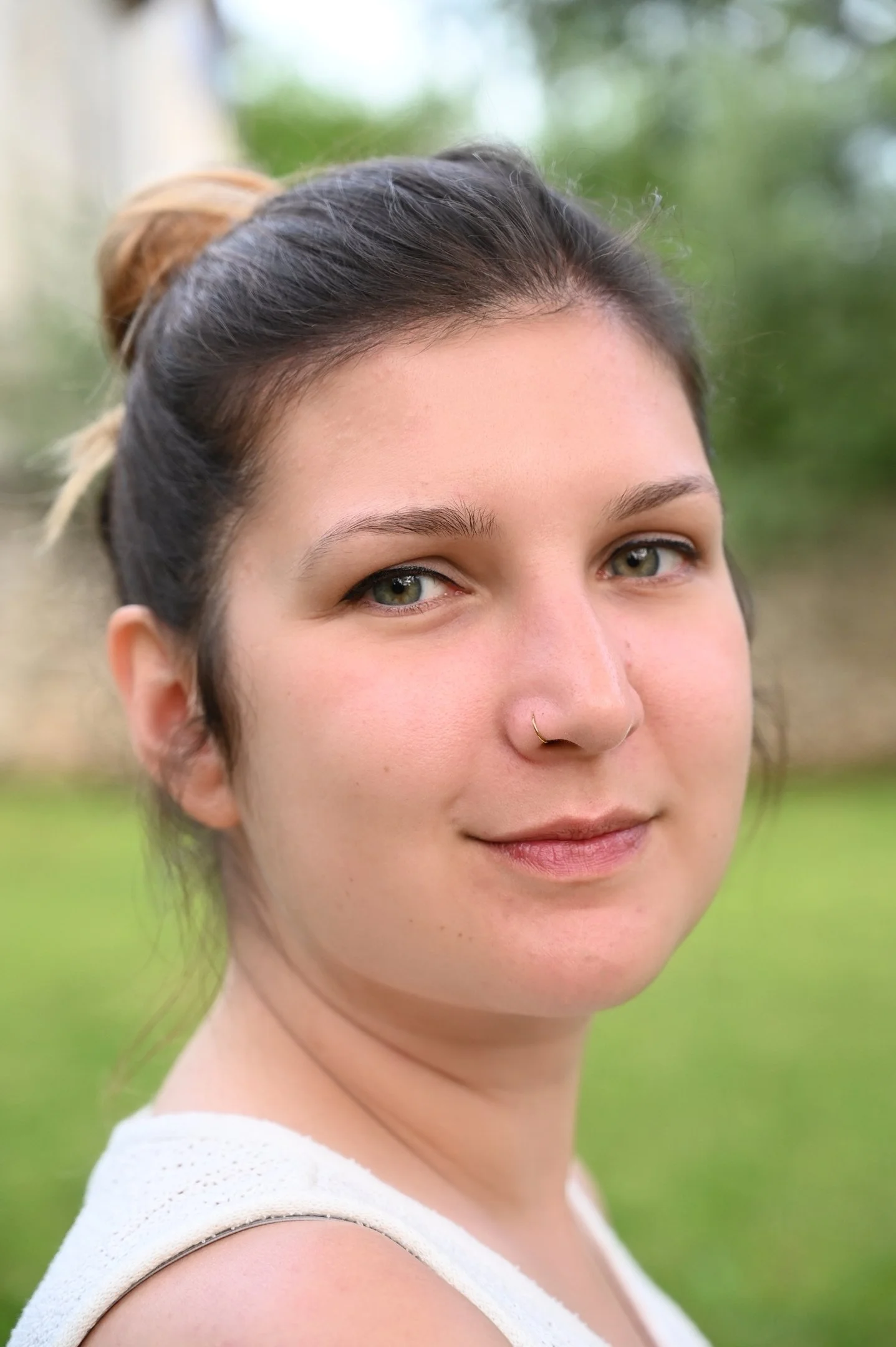 Nahe Portrait einer jungen Frau mit dunklem Haar, die einen Nasenring trägt, lächelnd im Freien bei bewachsenen Hintergrund.