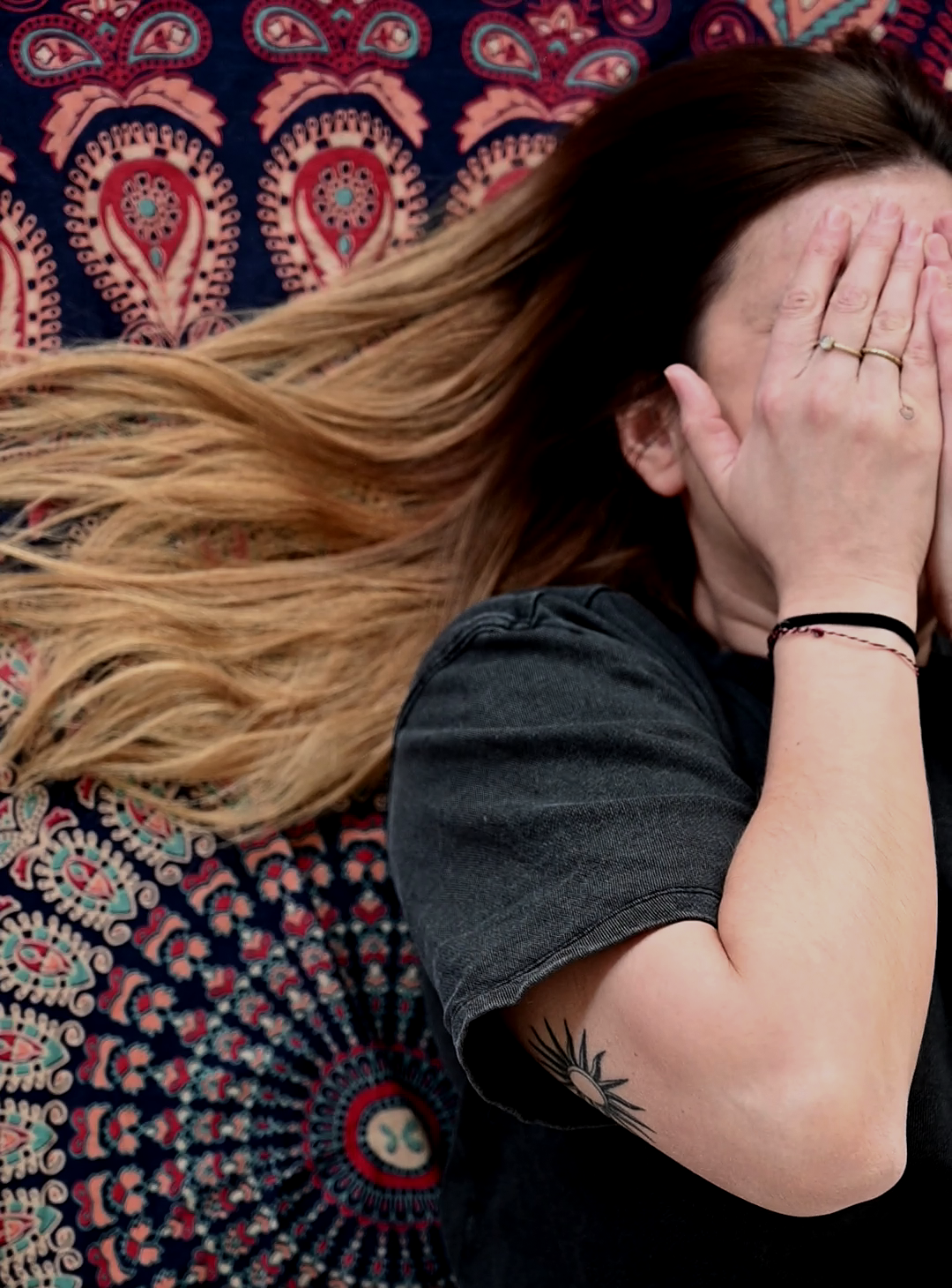 Eine Person mit langen, braunen Haaren, die ihr Gesicht mit den Händen bedeckt, liegt auf einem bunten, mandalaartigen Teppich.