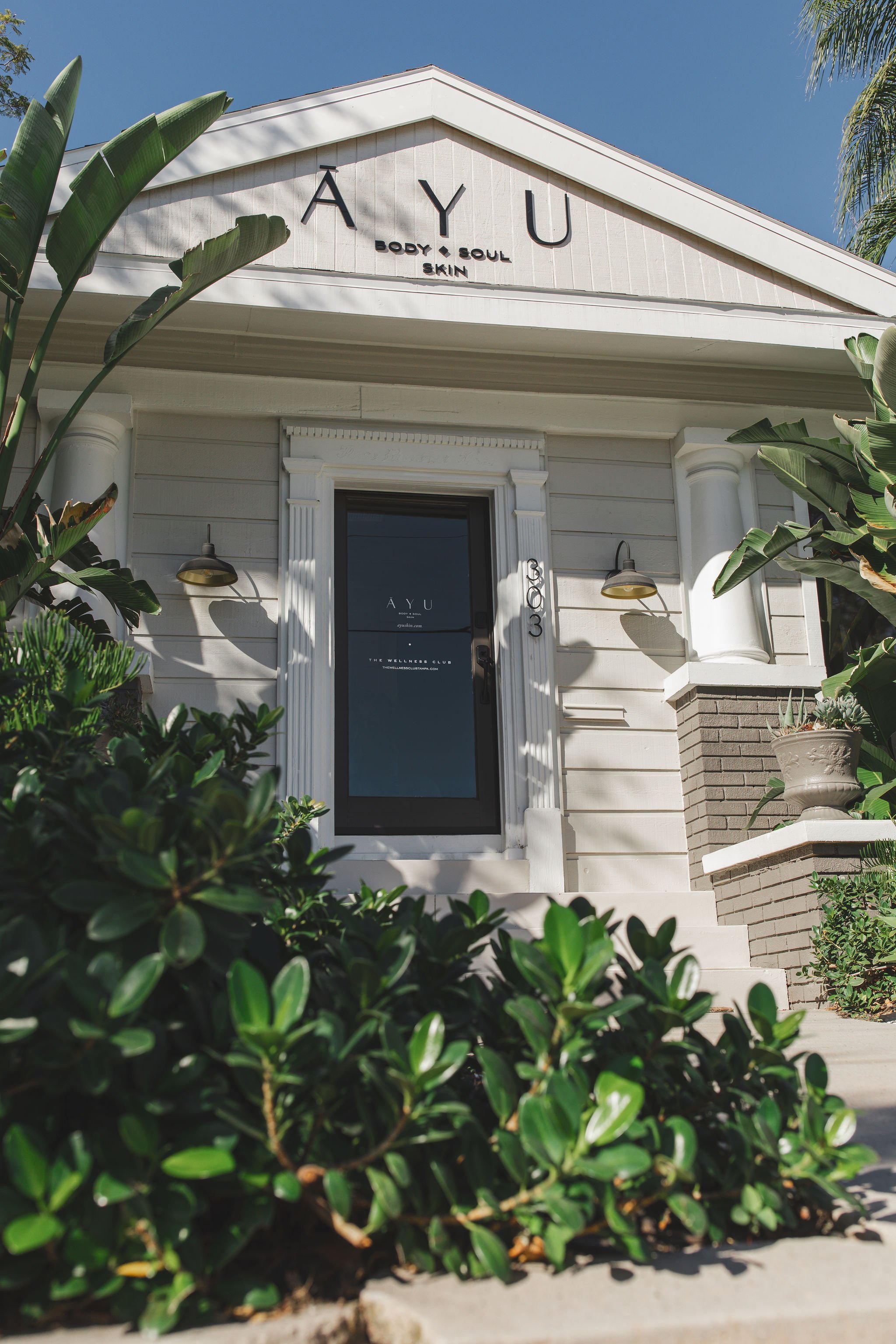 White building with a sign that reads 'AYU Body & Soul Skin,' surrounded by green plants and bushes, with a blue sky in the background.