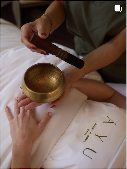 Person holding a singing bowl and a mallet, preparing to play it while lying on a bed with white sheets that have the words 'A Y U' embroidered on them.