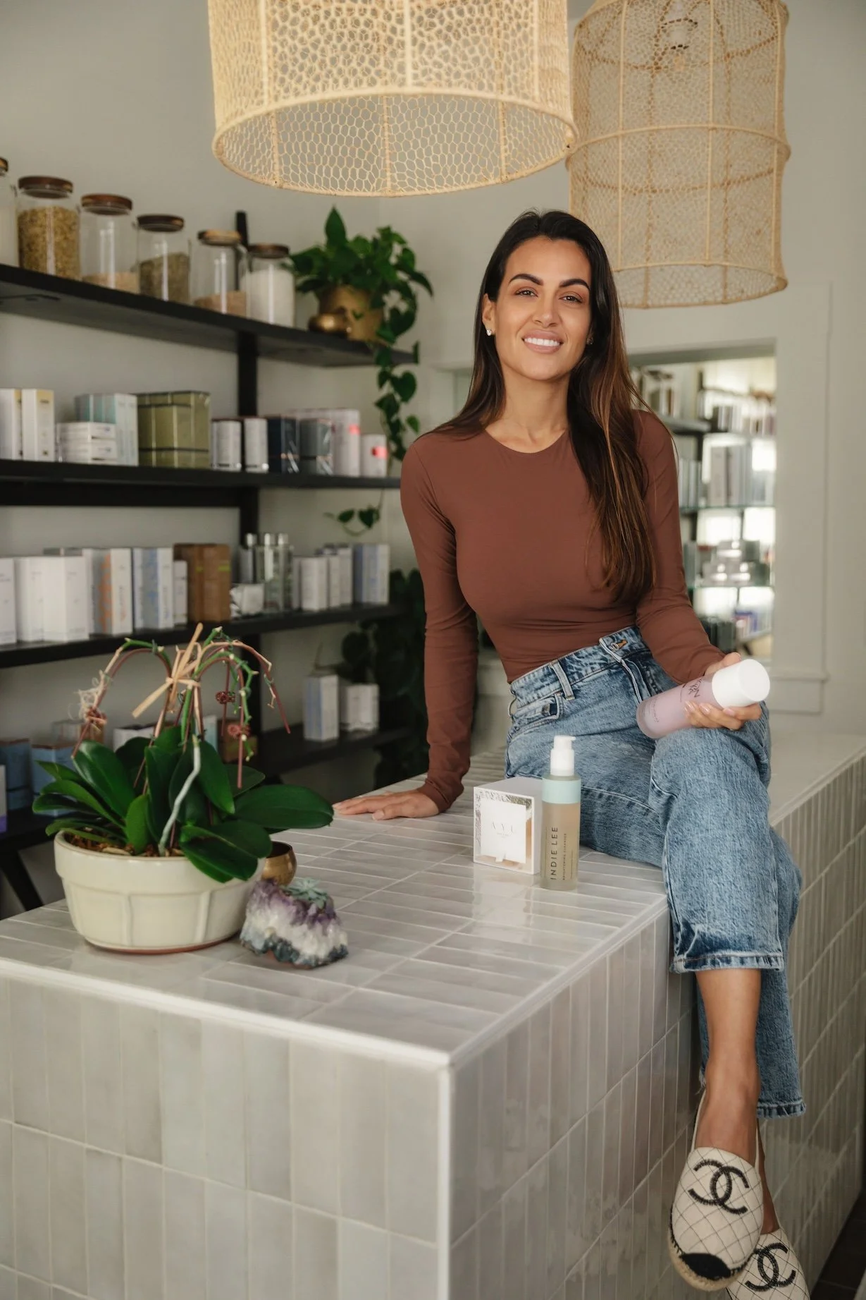 Ayu skin's founder Alex is holding a bottle of skincare product in her hand and smiling at the camera. The background features shelves with jars, books, and a green plant, along with large decorative hanging lights.