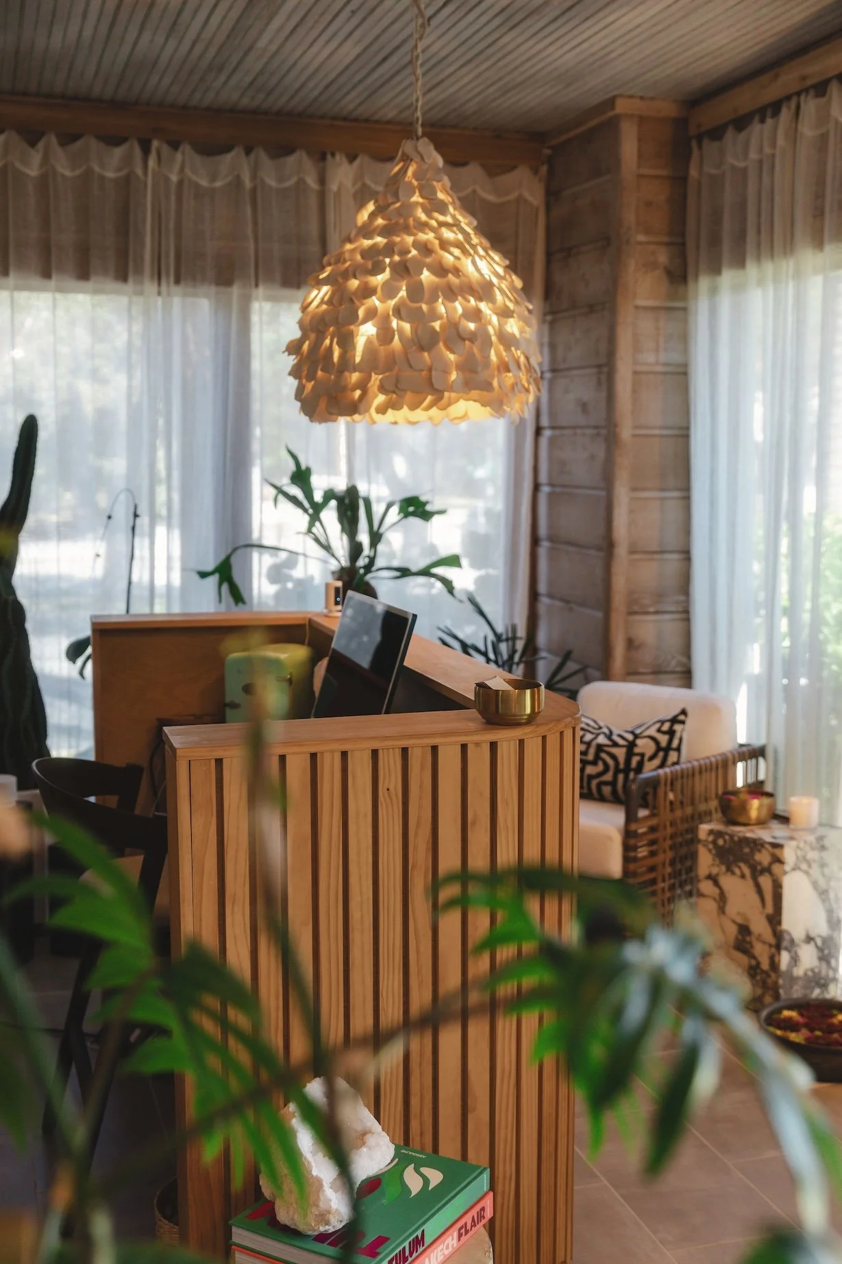 Interior of a cozy living space with large windows, sheer curtains, and a wooden ceiling. A stylish pendant light made of seashells hangs from the ceiling. There are several plants, a wooden divider, a computer monitor, a sofa with patterned cushions, and decorative items including bowls and candles.