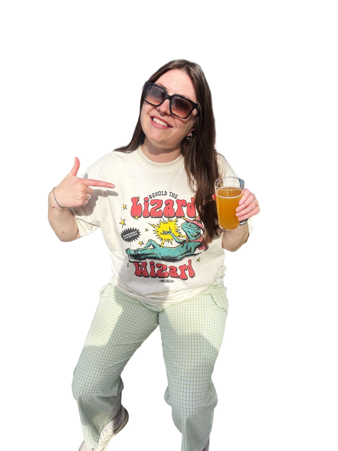 A woman wearing dark sunglasses and a graphic T-shirt smiling and pointing at herself while holding a beer. She is dressed in plaid pants and sneakers, with an energetic pose.
