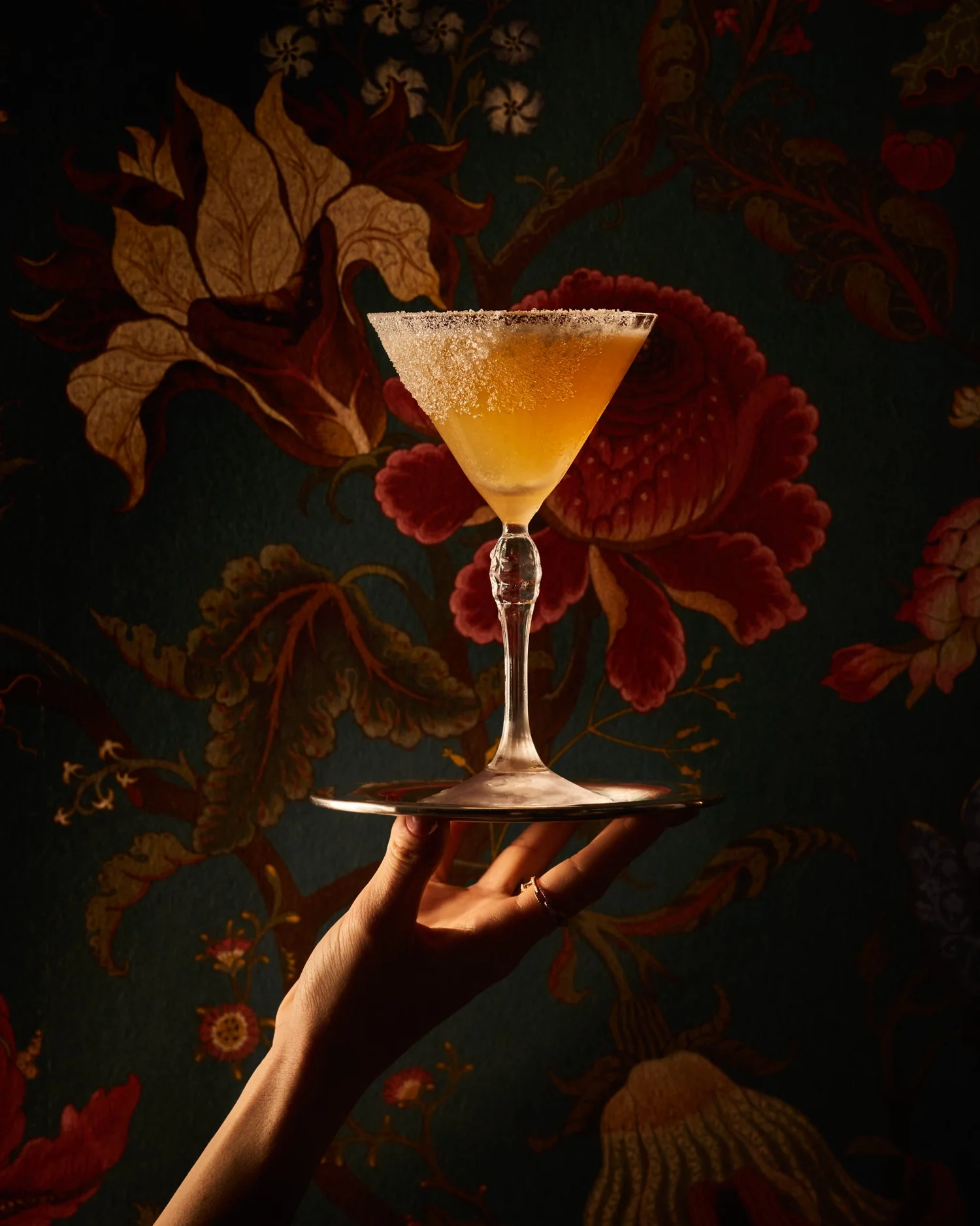 A hand holding an elegant cocktail in front of Left Door’s floral wallpaper