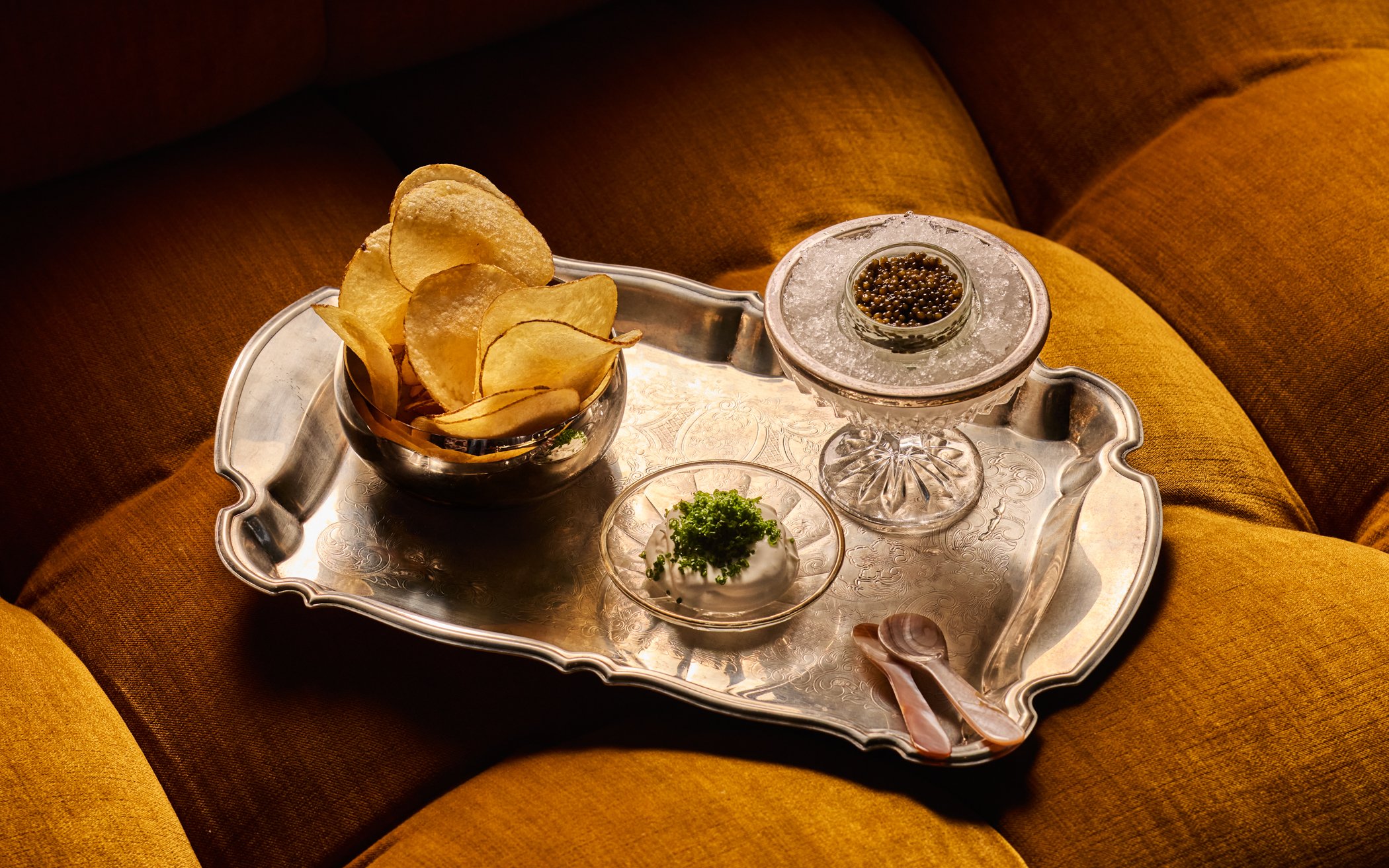 Caviar in tin in glass filled with ice with sides of cream and chips on tray on a yellow couch. . Photo credit: Nicola Parisi