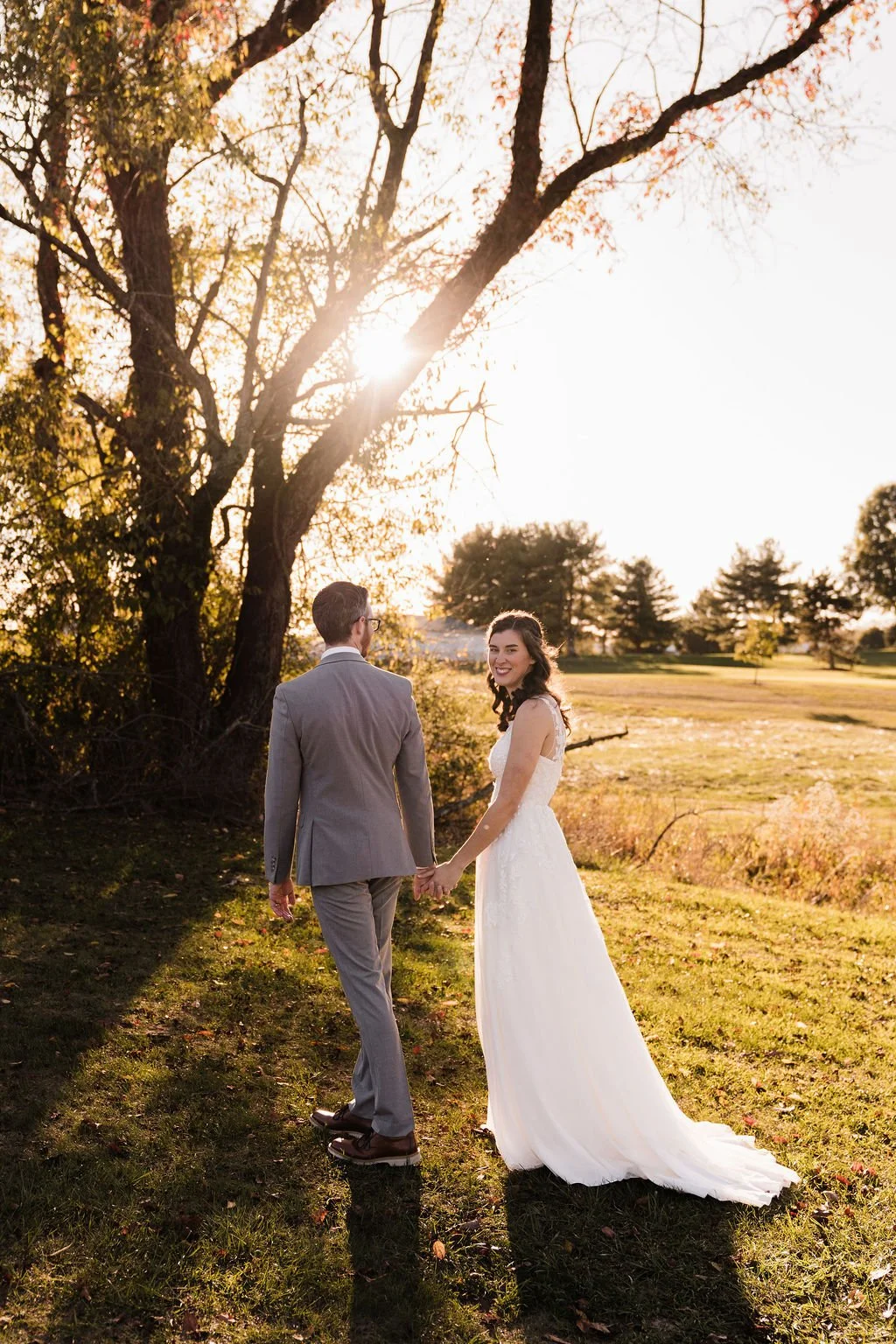 A woman in a white dress looks over her shoulder as she holds hands and walks with man in gray suit