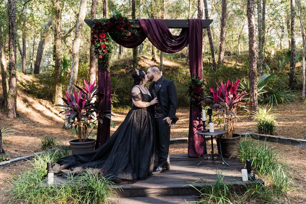 Bride and groom both wearing black wedding attire share a kiss underneath a wedding arch covered in red drapes at outdoor forest wedding ceremony