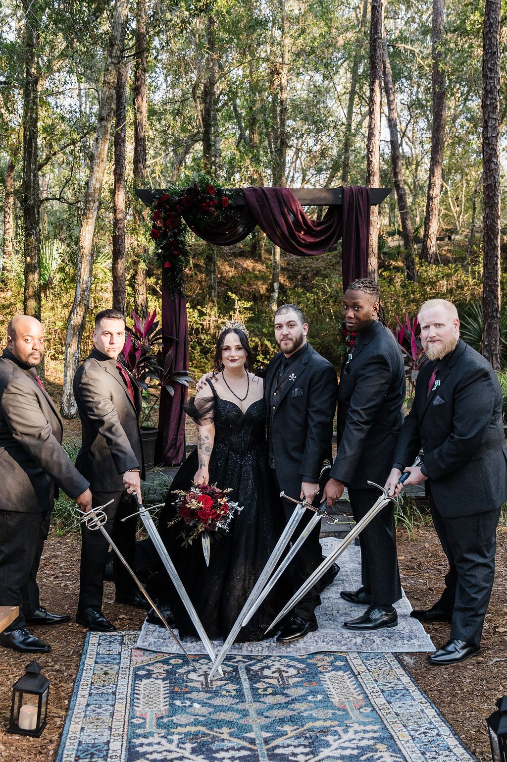 Bride in black sparkling wedding dress and groom in black wedding attire surrounded by groomsmen in black attire all holding swords and daggers pointing at the ground
