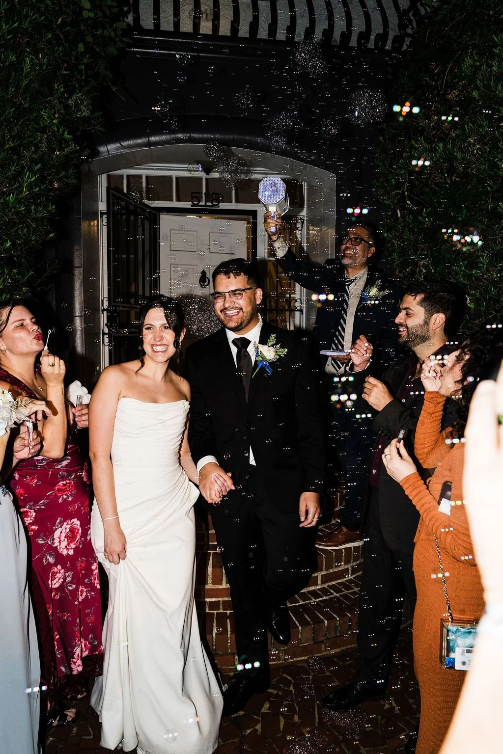 Nighttime photo of bride in white strapless dress holding hands with groom in black suit as wedding guests blow bubbles on either side of them