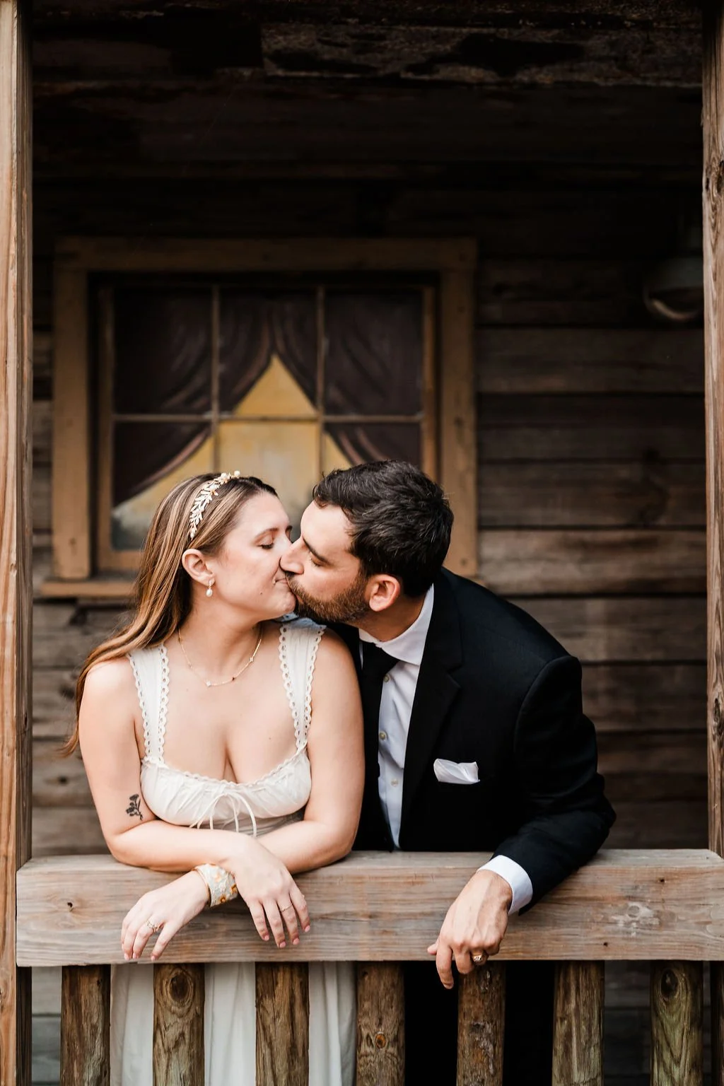 Bride in white cotton dress and metal wreath headband kissing groom in black suit on wood porch of wood building