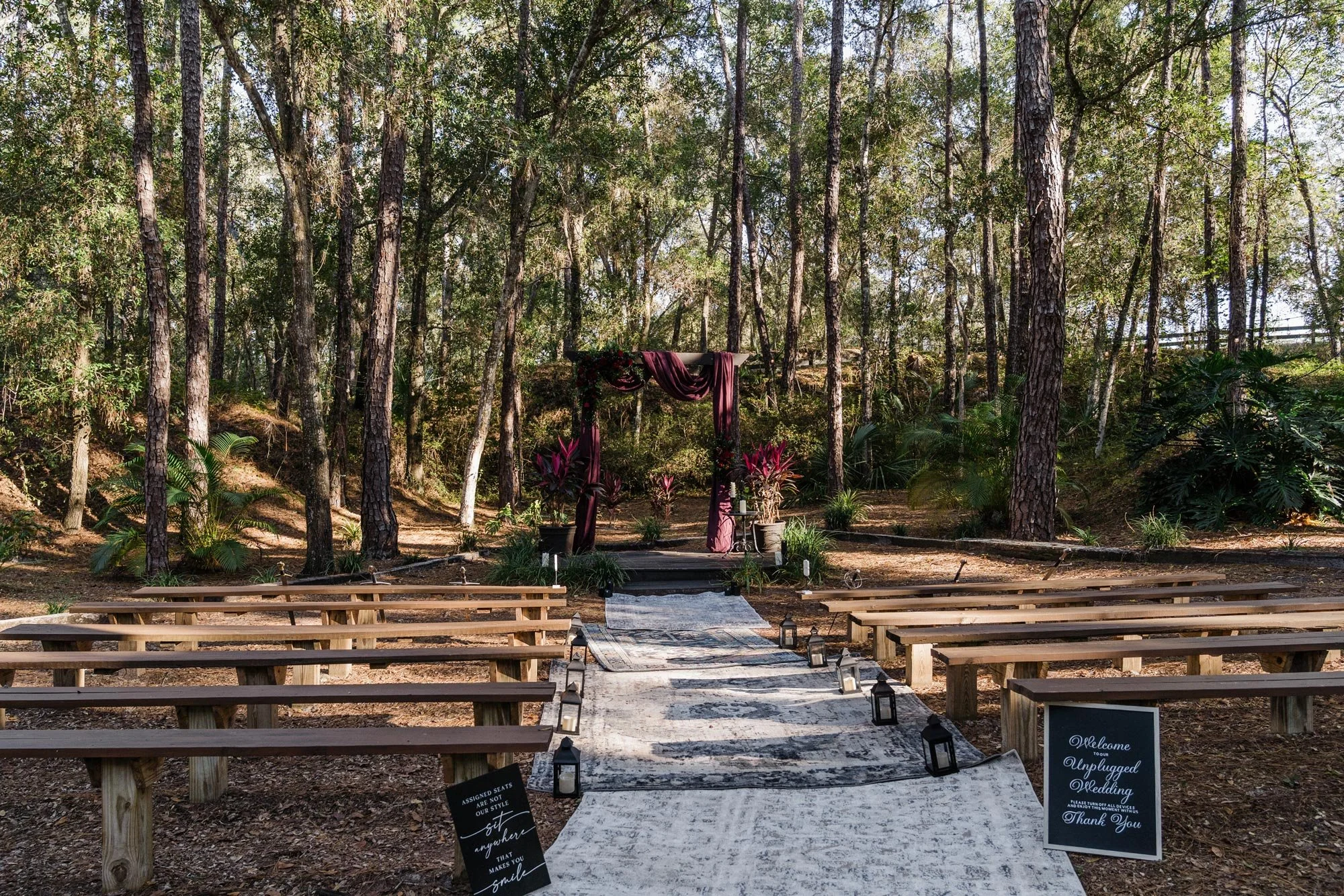 Wooden arch with red draped fabric set up for a wedding in woods with blue carpets lining the walkway and wooden bench seating