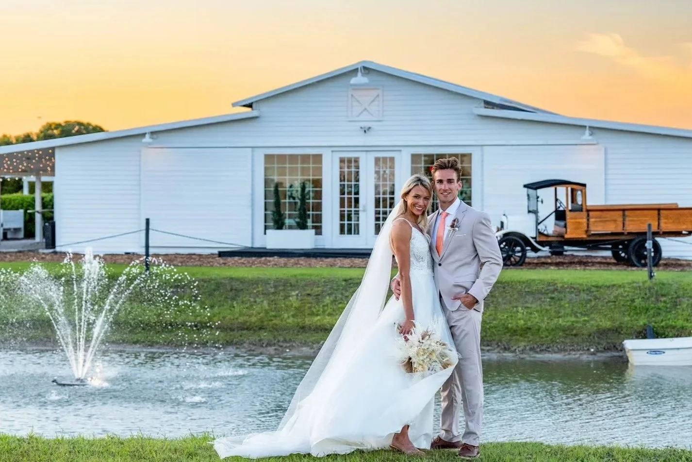 Bride in white dress and groom in gray suit pose in front of lake with fountain and white barn at sunset