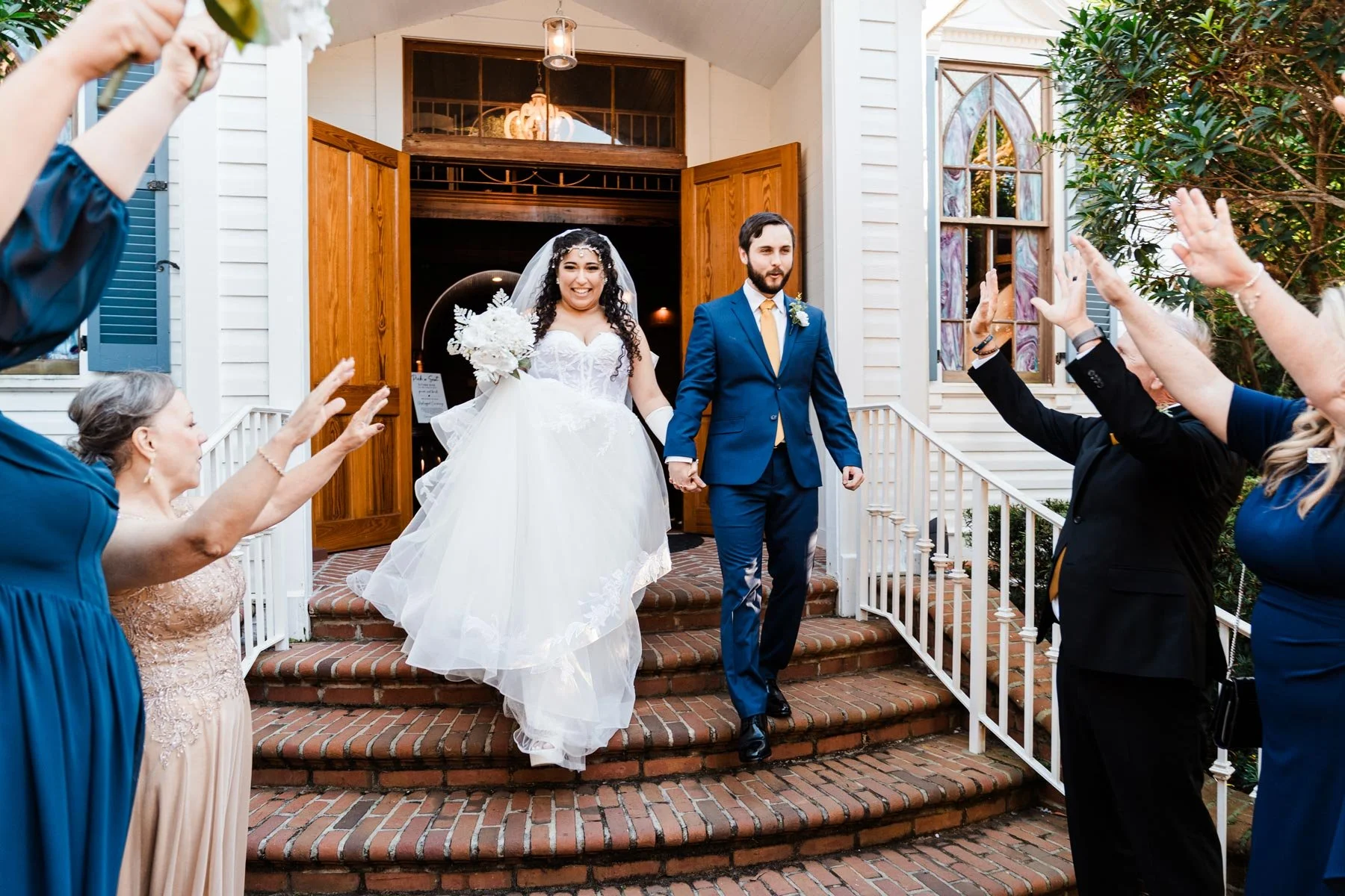 Bride in white lacy dress and groom in blue suit hold hands and walk down brick steps of white chapel with wood doors