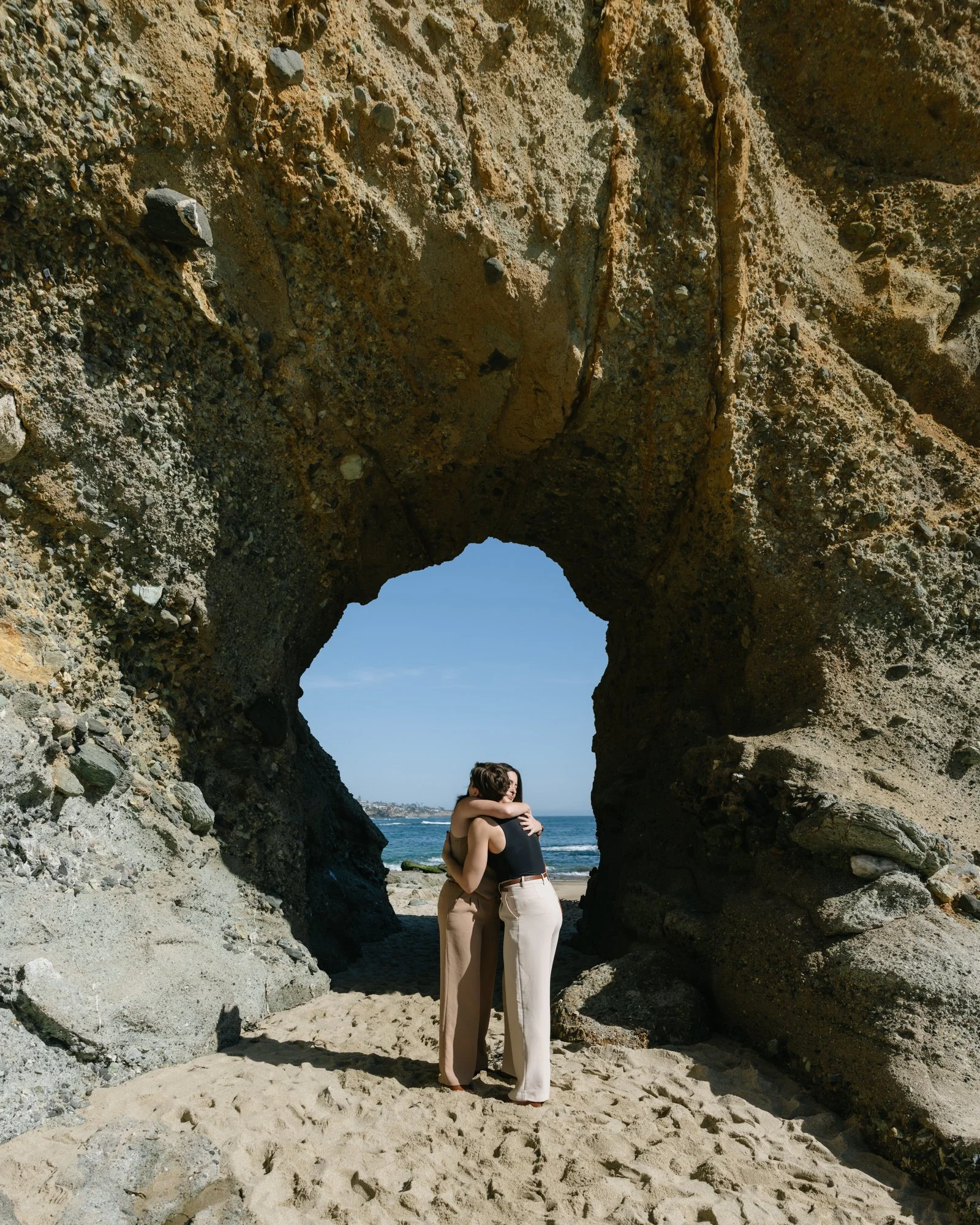 A couple embracing under a large natural rock arch formation on a beach, with the ocean and blue sky in the background.