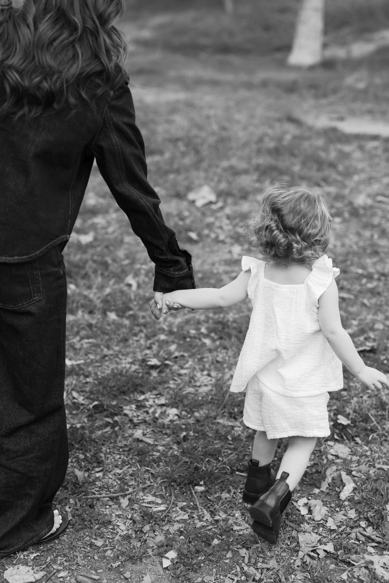 A woman and a young girl holding hands while walking outdoors on a leaf-covered ground, with trees in the background, in black and white.