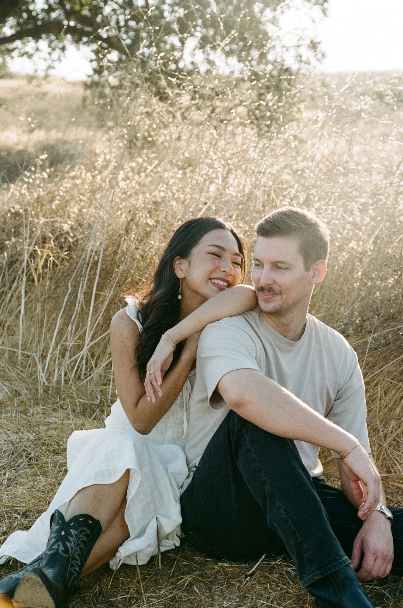A young woman and a young man sitting together in a field of dry grass, smiling and enjoying each other's company during a sunny day.