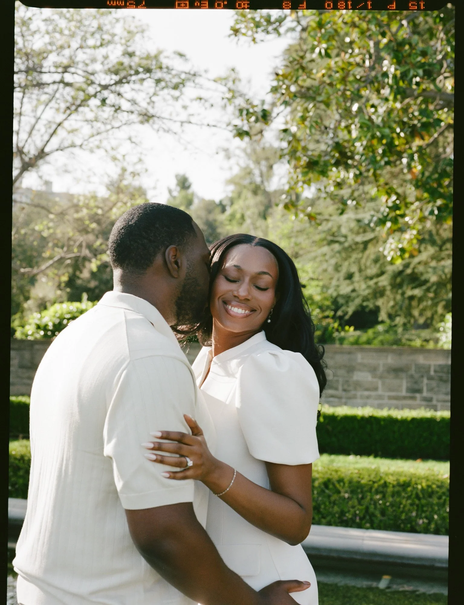 A couple embracing outdoors, with greenery and trees in the background, both dressed in white and smiling.