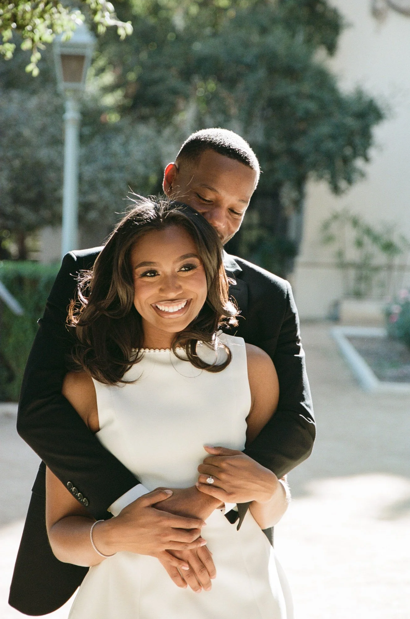 A smiling bride in a white dress with a diamond ring, embraced by a groom in a black suit, outdoors with trees and sunlight.
