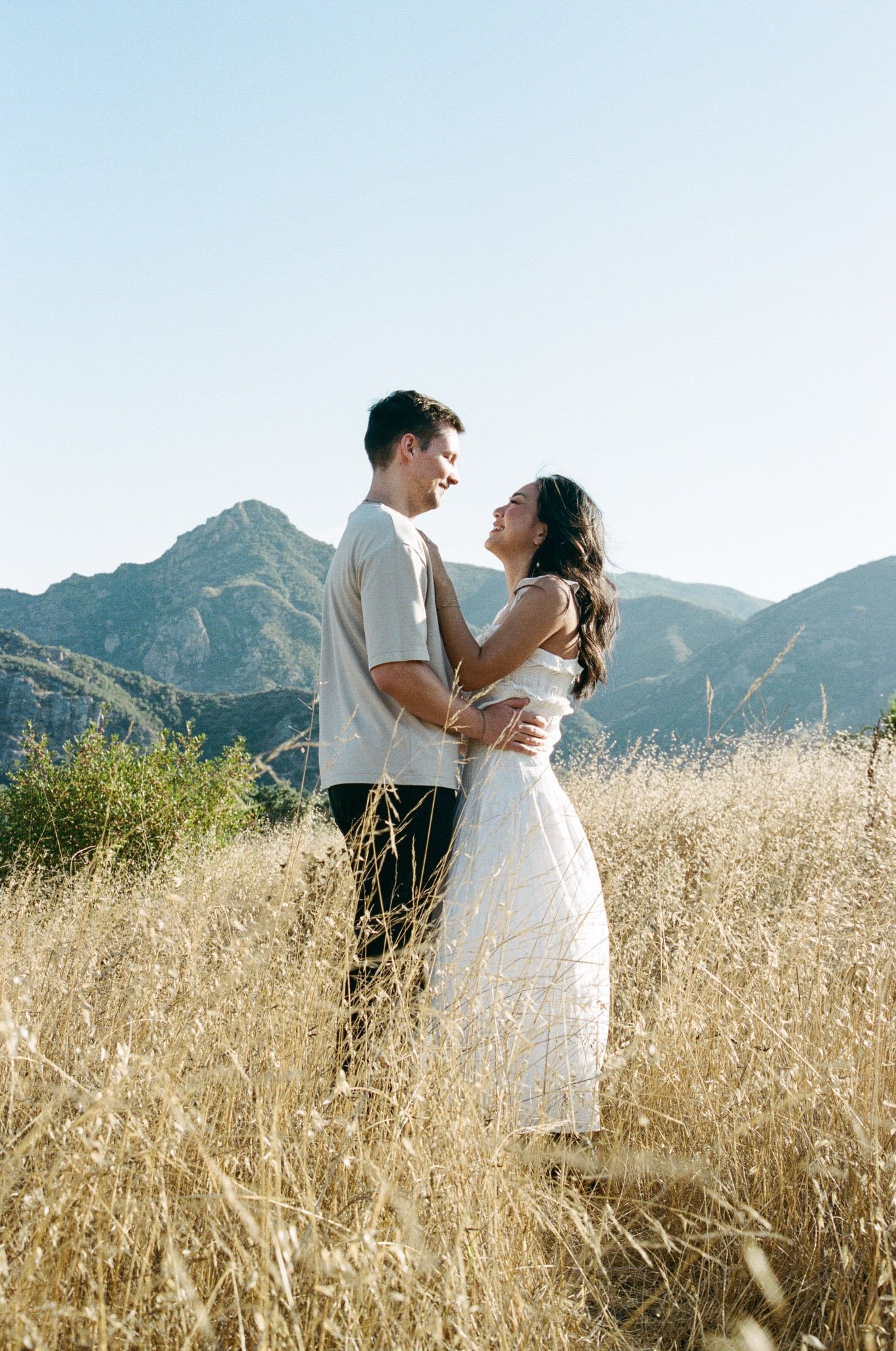 A couple standing close together in a field of tall, yellow grass with mountains in the background, enjoying a bright and clear day.