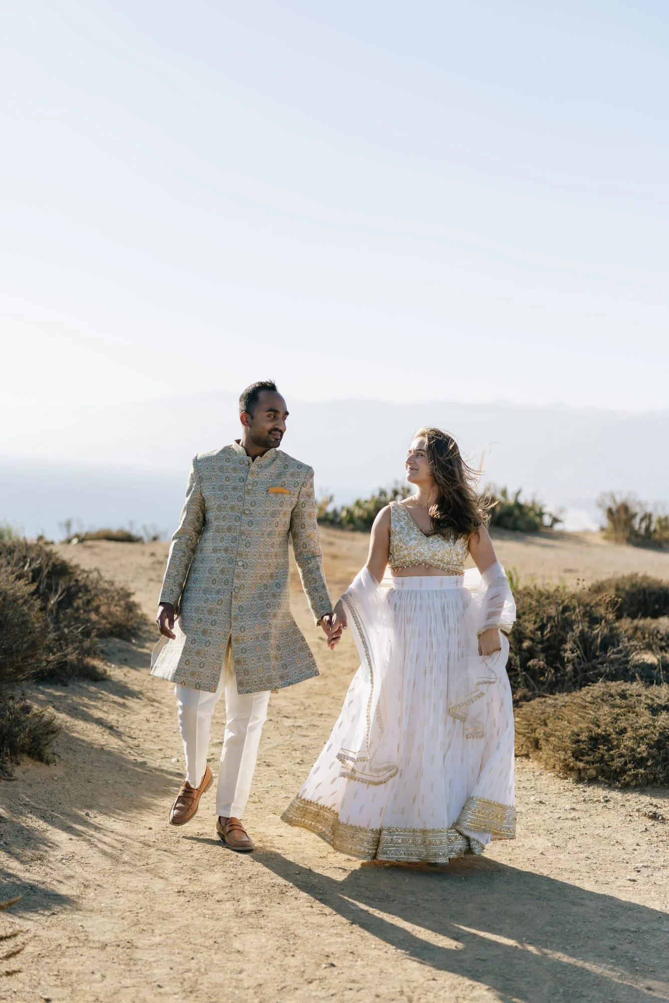A couple in traditional Indian attire holding hands and walking outdoors on a dirt path with distant hills and sky in the background.