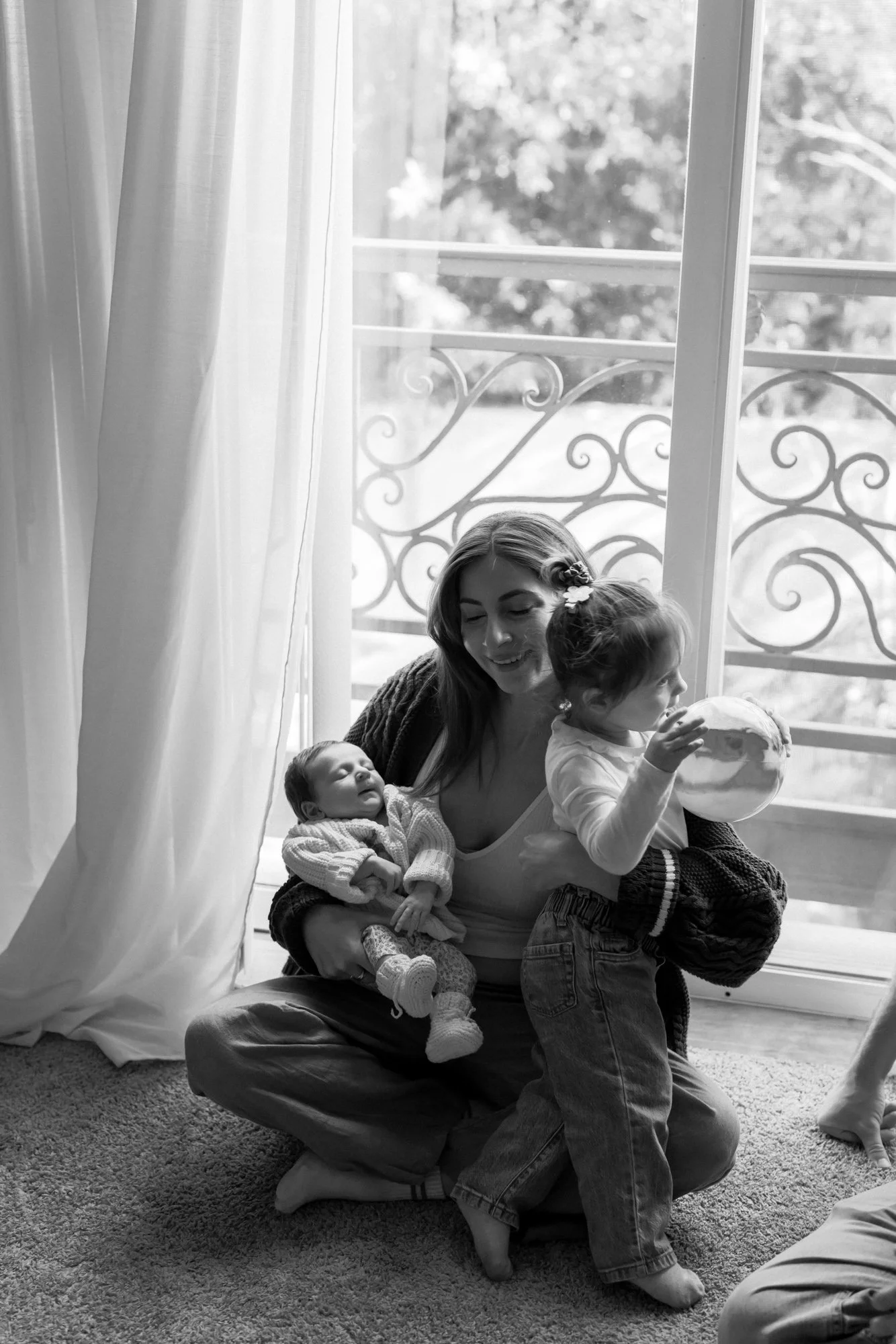 A woman sitting on her knees on a carpeted floor, holding two children, one in each arm. One child is a baby being held in her left arm, and the other is a young girl standing on her knee, holding a ball. They are near a large window with curtains pa