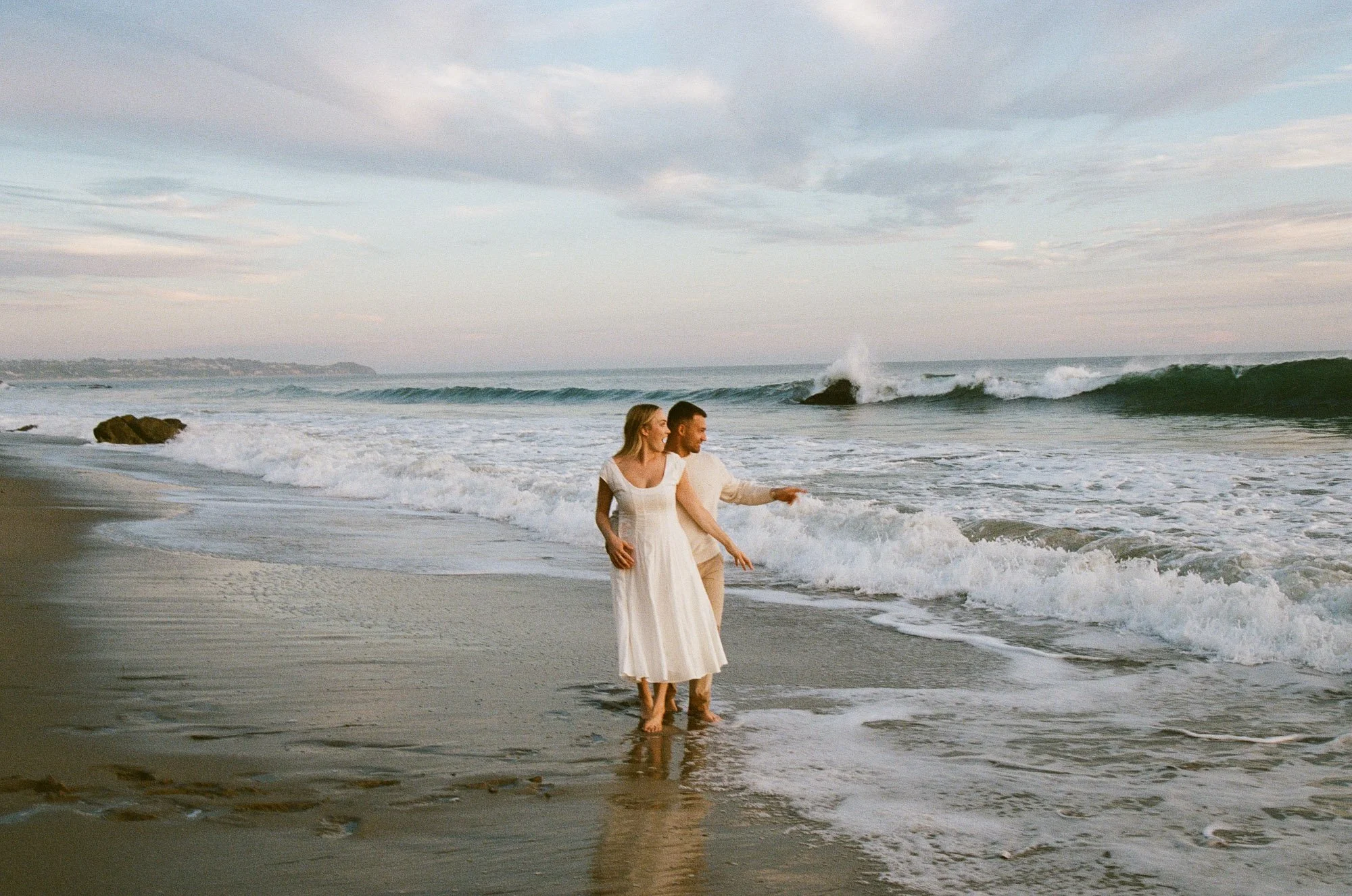A couple in wedding attire enjoying a walk on the beach, with waves crashing in the background under a cloudy sky.