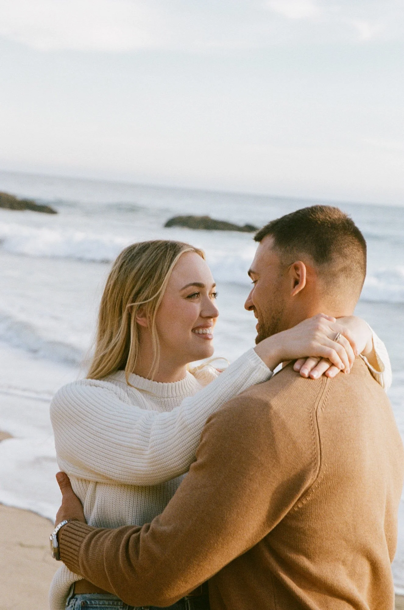 A smiling woman and a man hold each other close on a beach with waves and rocks in the background.