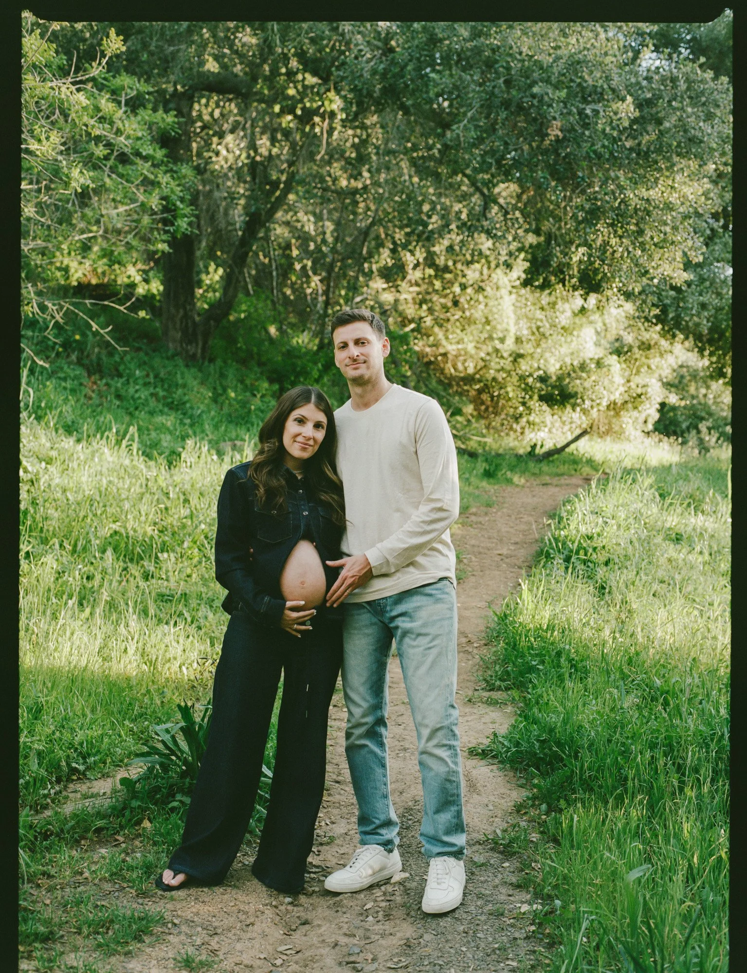 A pregnant woman and a man standing on a dirt path in a lush green park with trees and grass. The woman is cradling her belly, and the man is standing next to her with one hand on her belly.