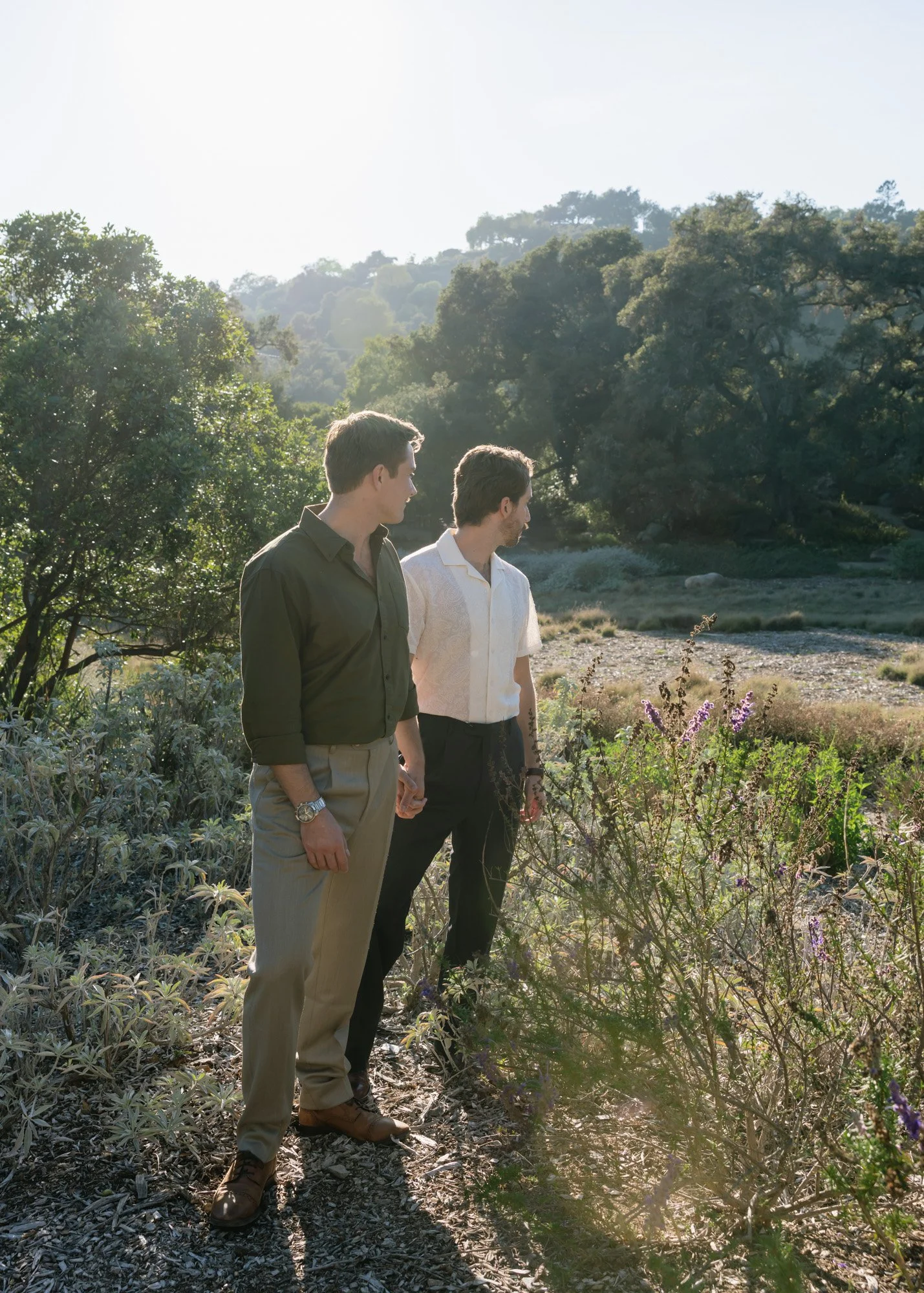 Two men holding hands and looking at plants in a lush outdoor garden or park with trees and hills in the background during the day.