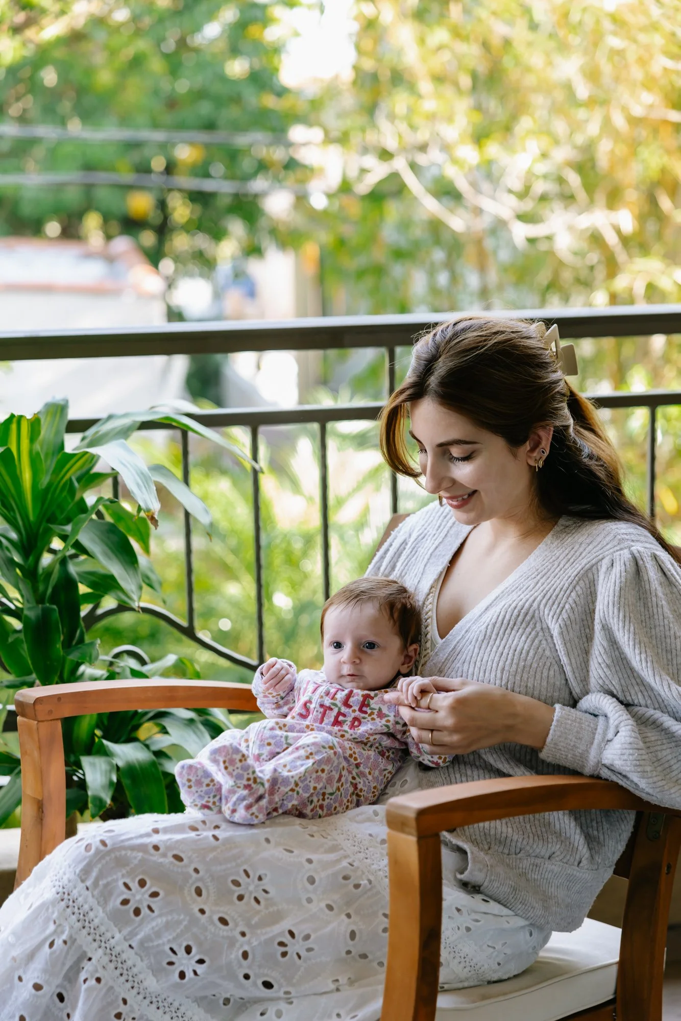 A woman with brown hair and earrings sitting in a wooden chair with a baby girl on her lap, surrounded by indoor plants on a porch or balcony with a metal railing and greenery outside.