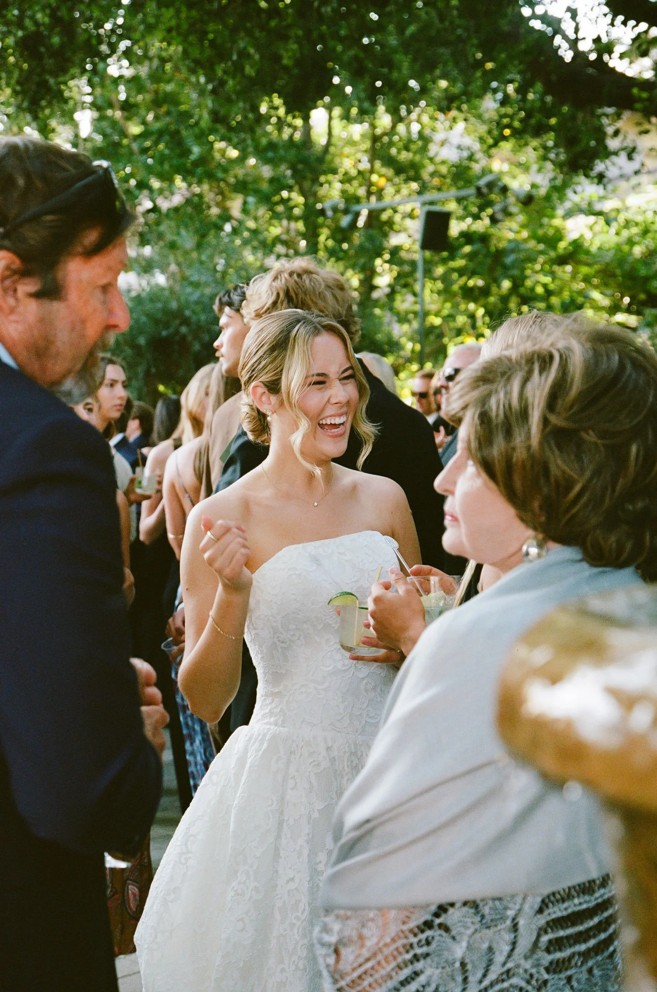 A woman in a white wedding dress laughing and holding a drink at an outdoor event with people around her.