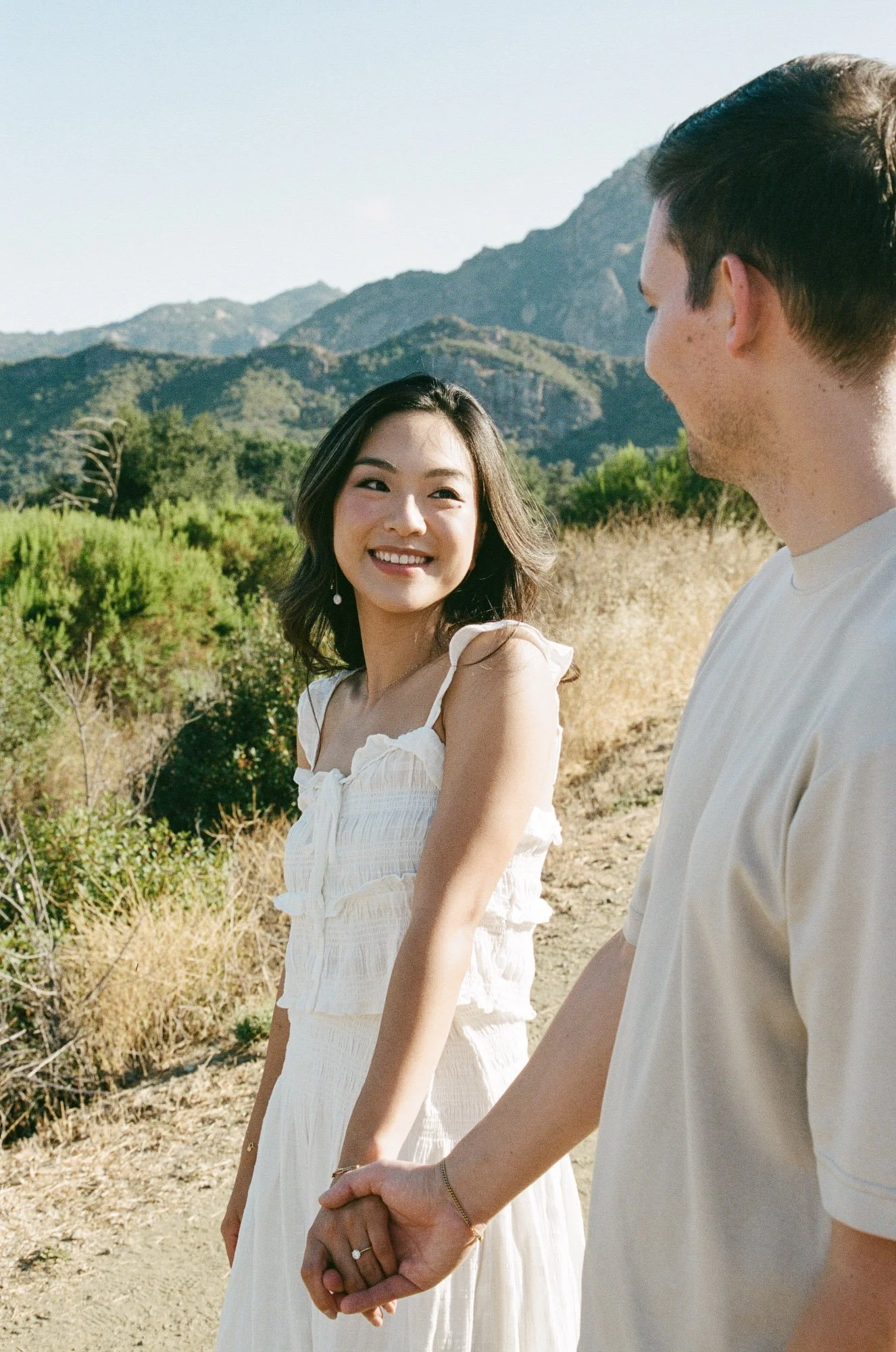 A young couple holding hands outdoors with mountains in the background. The woman is smiling and looking at the man, who is slightly turned away from the camera.