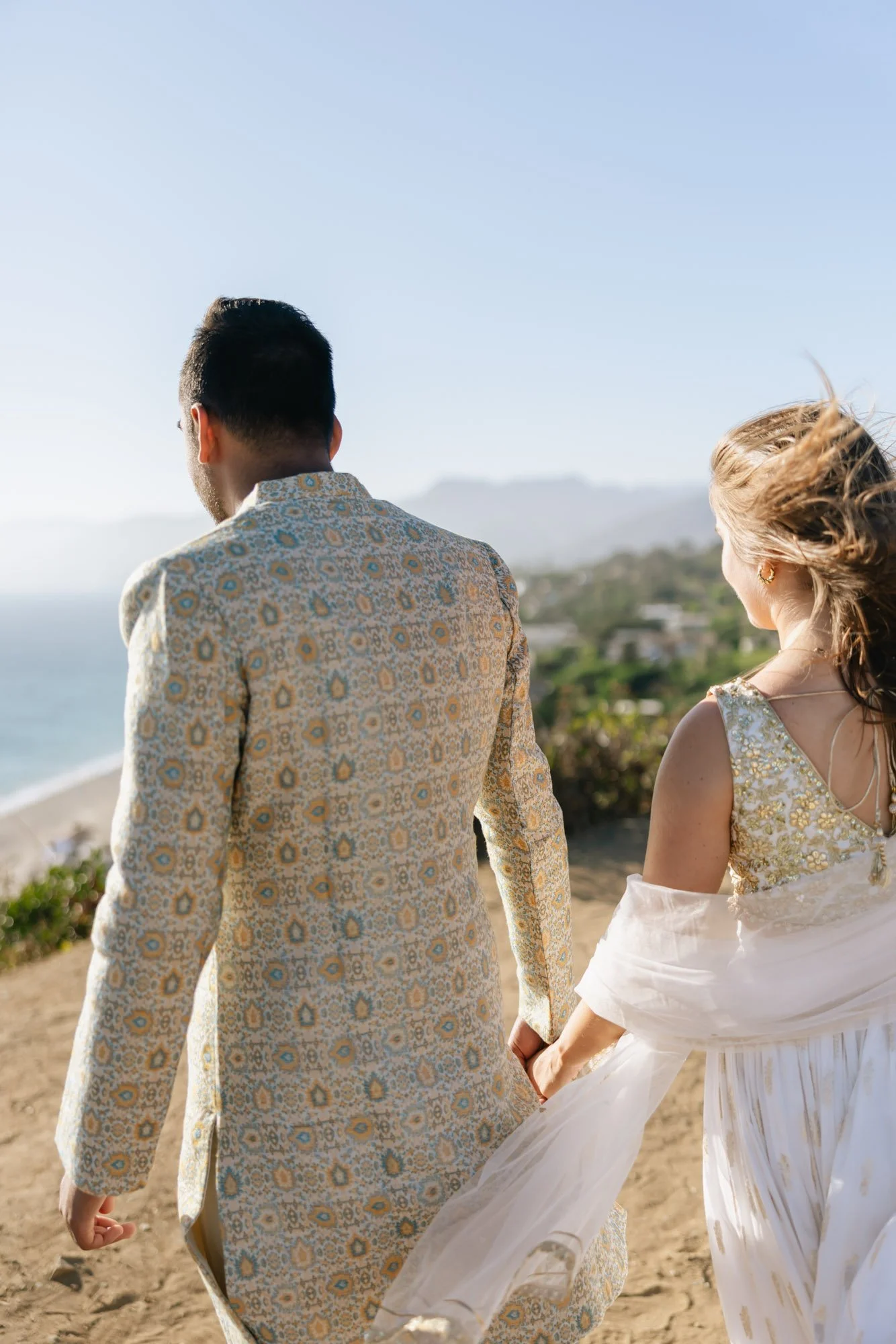 A man and a woman holding hands while walking on a beach. The man has short dark hair and wears a patterned beige and blue suit. The woman has curly blonde hair and wears a white and gold dress with a flowing skirt. In the background, there is a clea