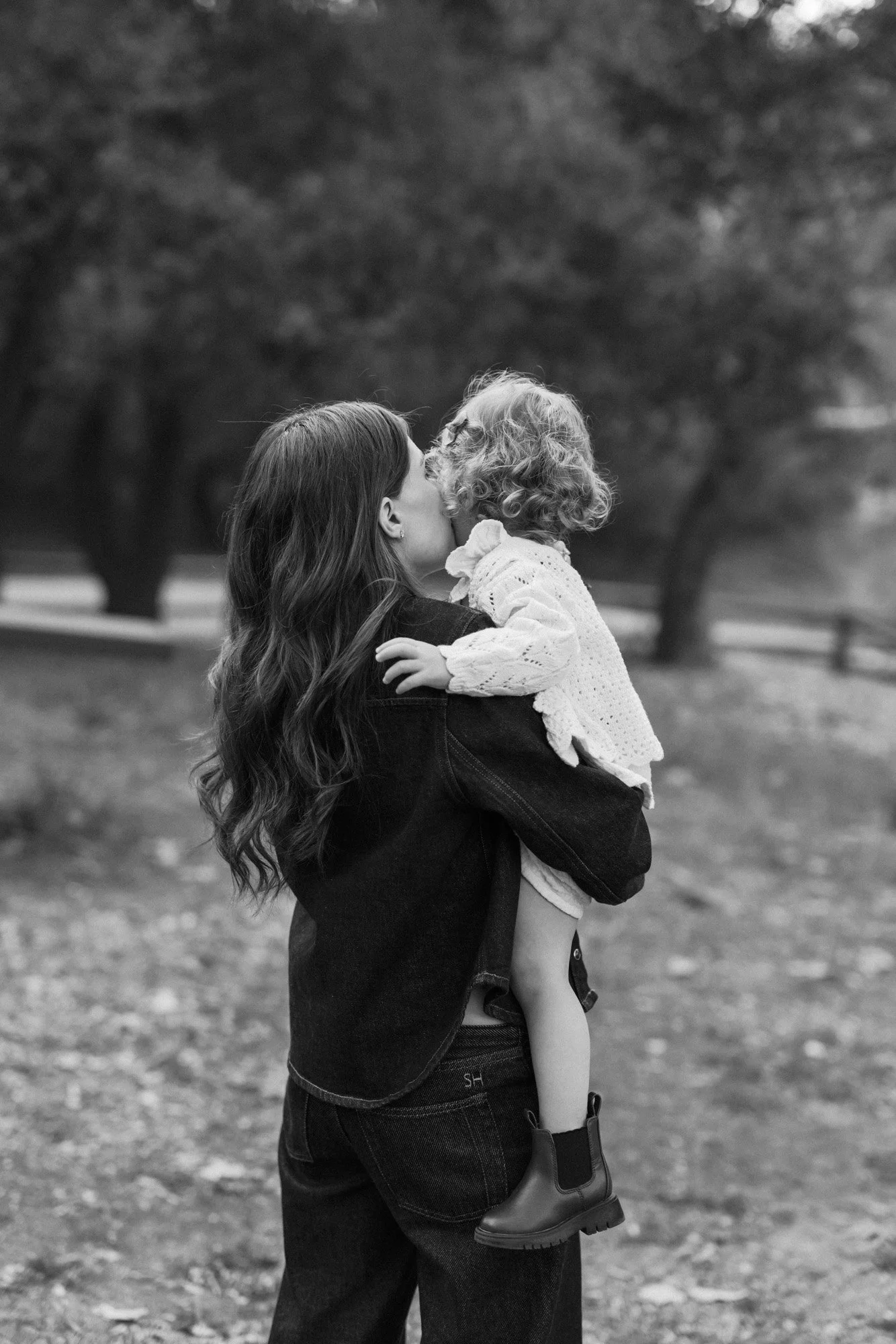 A woman with long wavy hair holding a young girl with curly hair and boots, kissing her on the lips outdoors in a park with trees in the background.