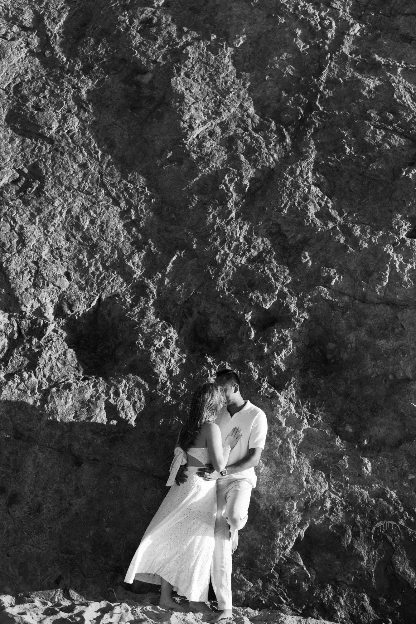 A couple is kissing in front of a rocky backdrop, with the woman wearing a white dress and the man dressed in white, both standing on sand.