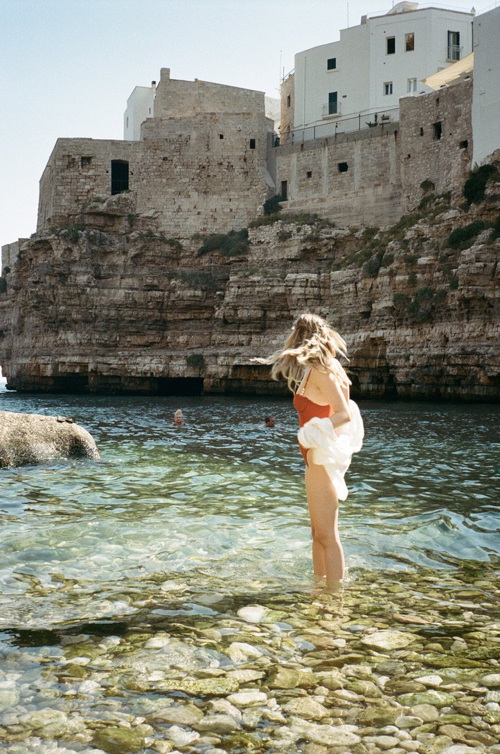 A woman standing in clear shallow water near rocky shoreline on a sunny day, with historic stone buildings on a cliff in the background.