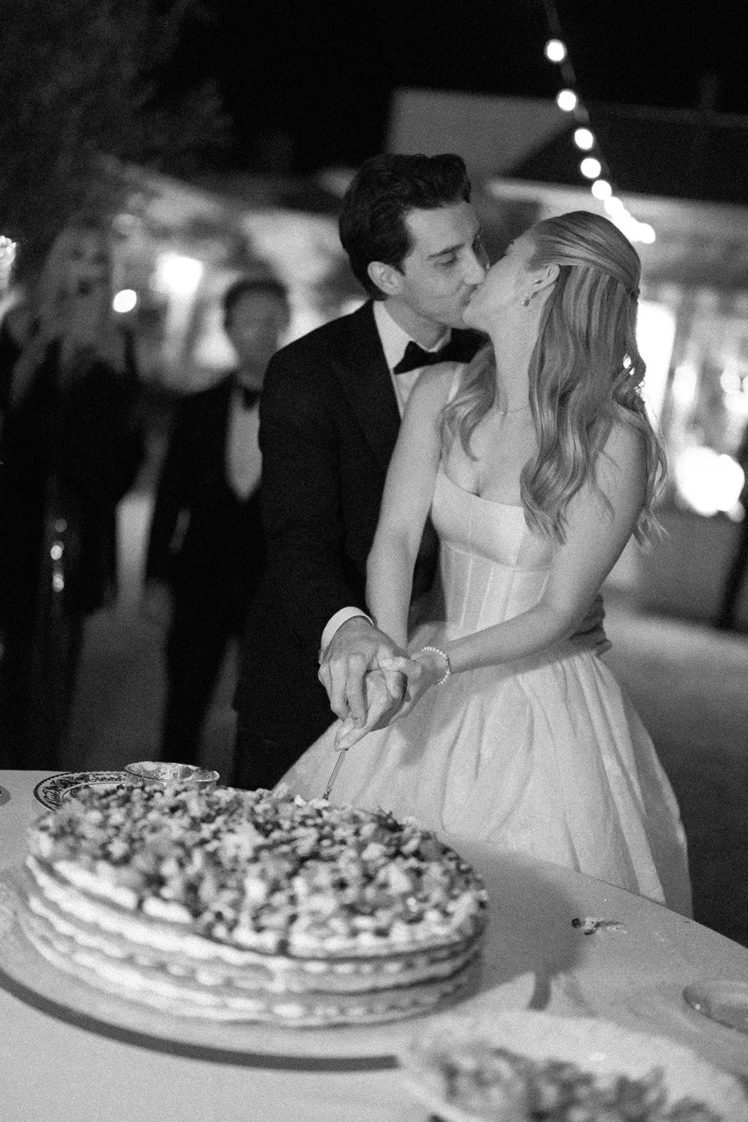A black-and-white photo of a couple kissing at a wedding reception, holding hands over a cake on a table, with blurred background of guests and string lights.